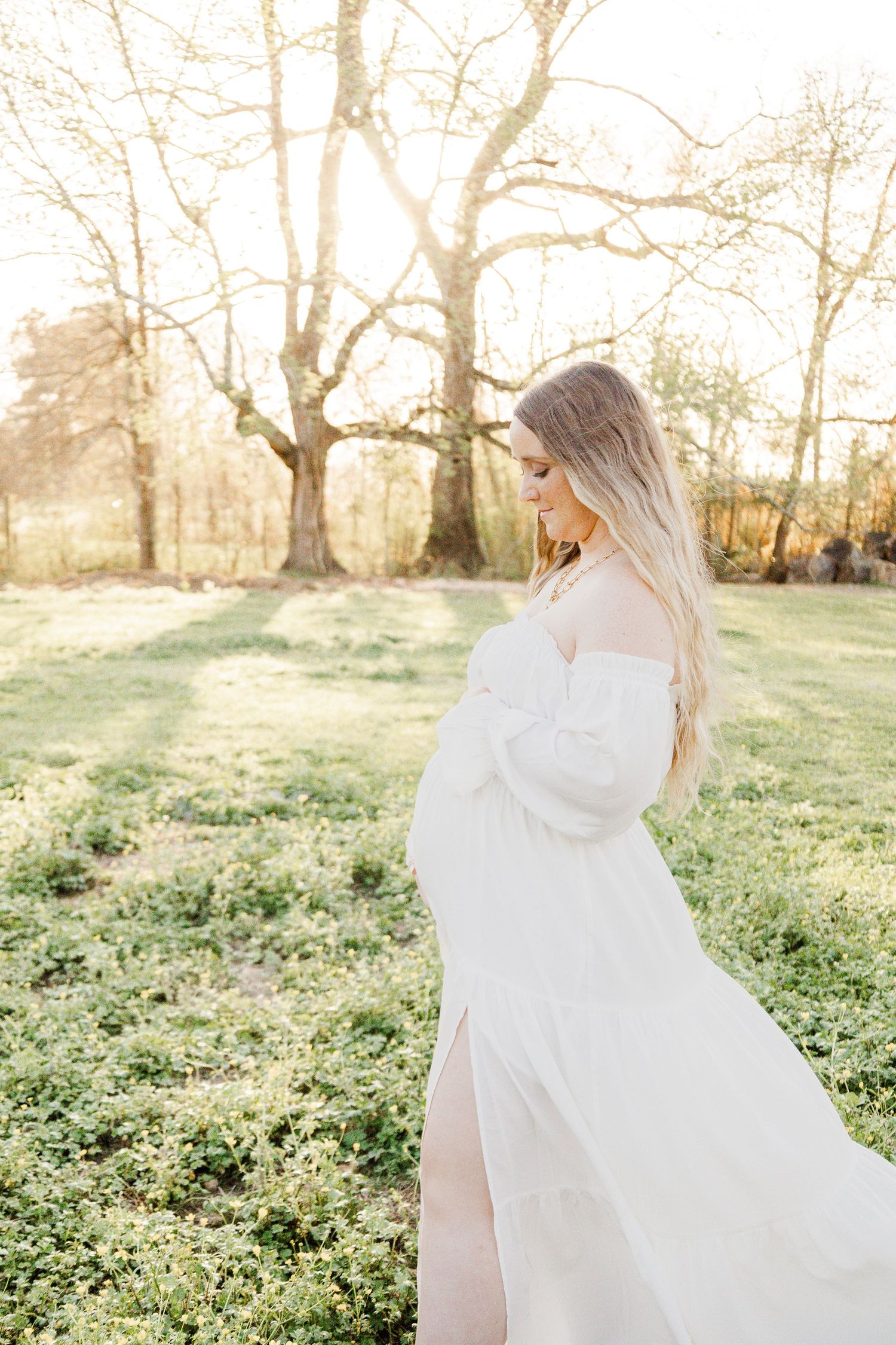 Pregnant woman in white off-shoulder dress standing in sunlit field with bare trees in background.