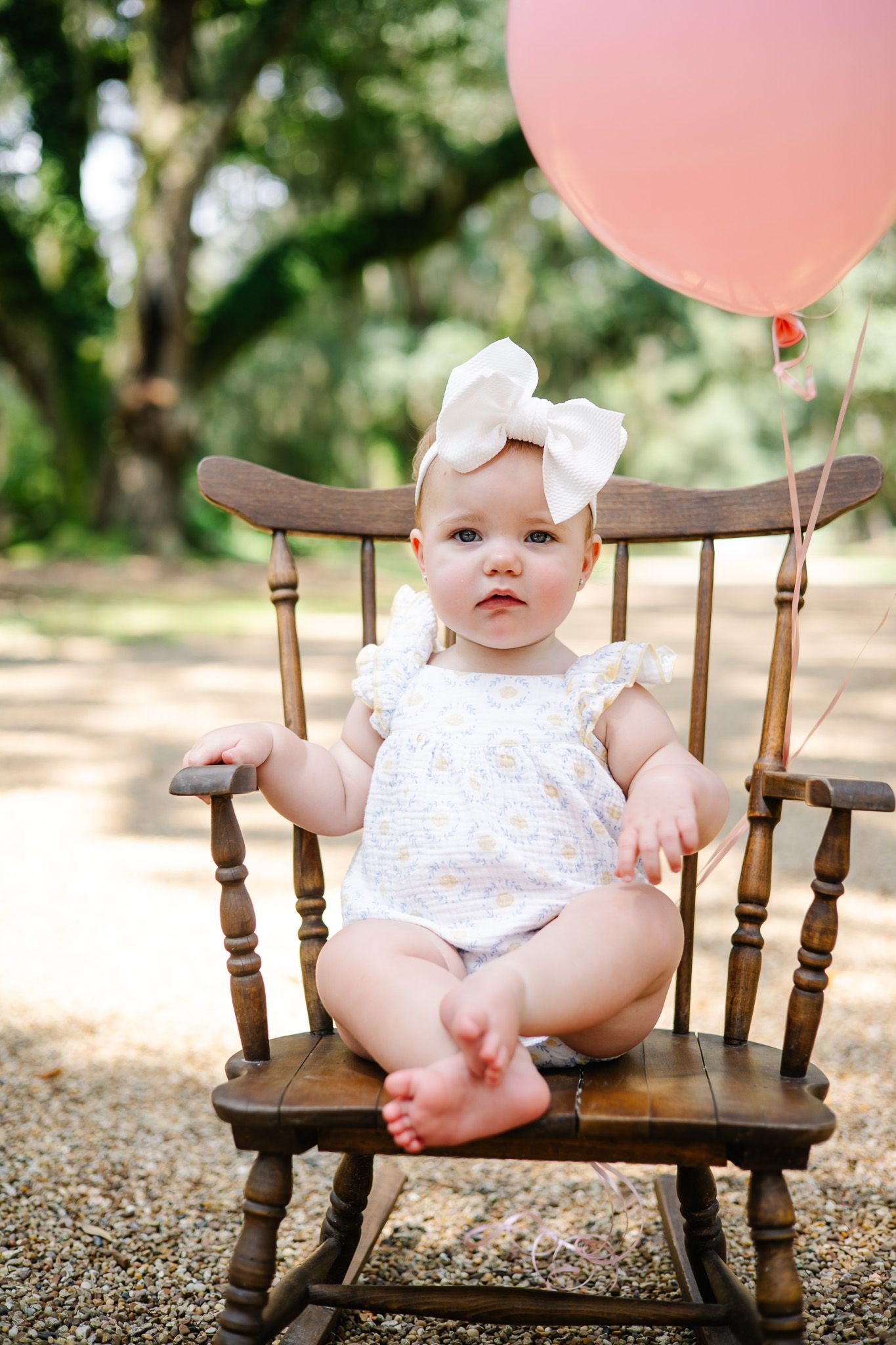 Baby girl in white floral romper and bow headband sits in wooden rocking chair with pink balloon outdoors.