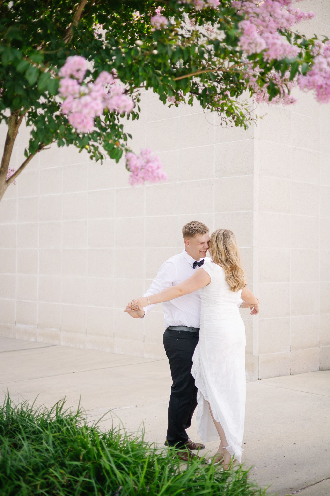 A couple shares a romantic dance and kiss beneath blooming pink crepe myrtle trees against a stone wall.