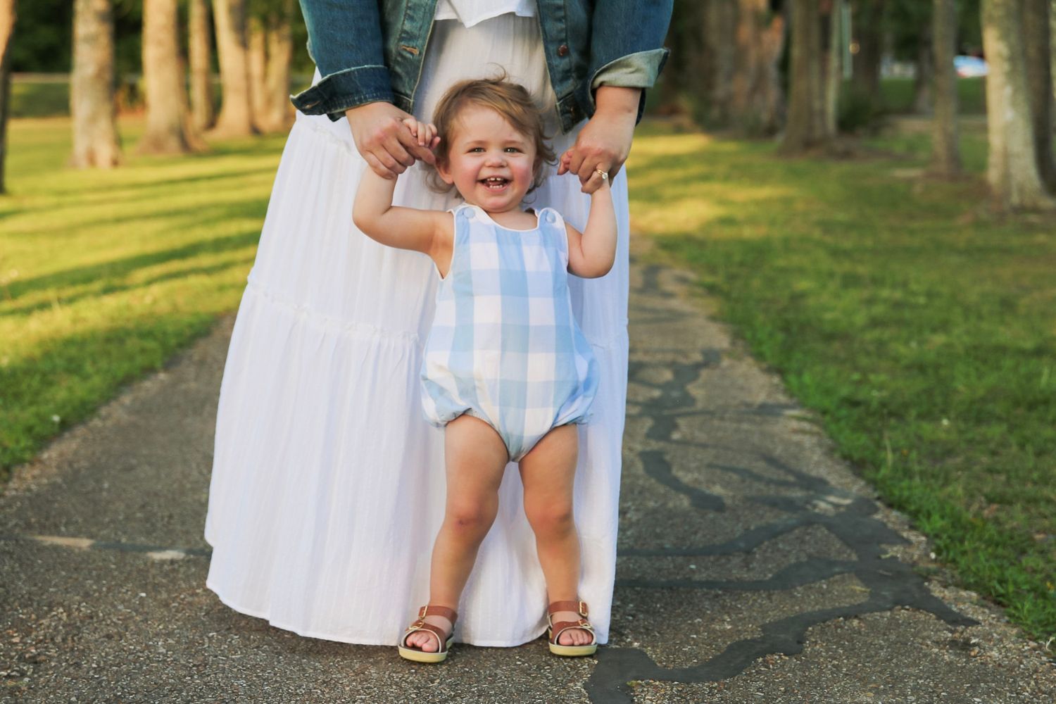 Mother holding toddlers hands helping baby stand on a paved path in a green park.