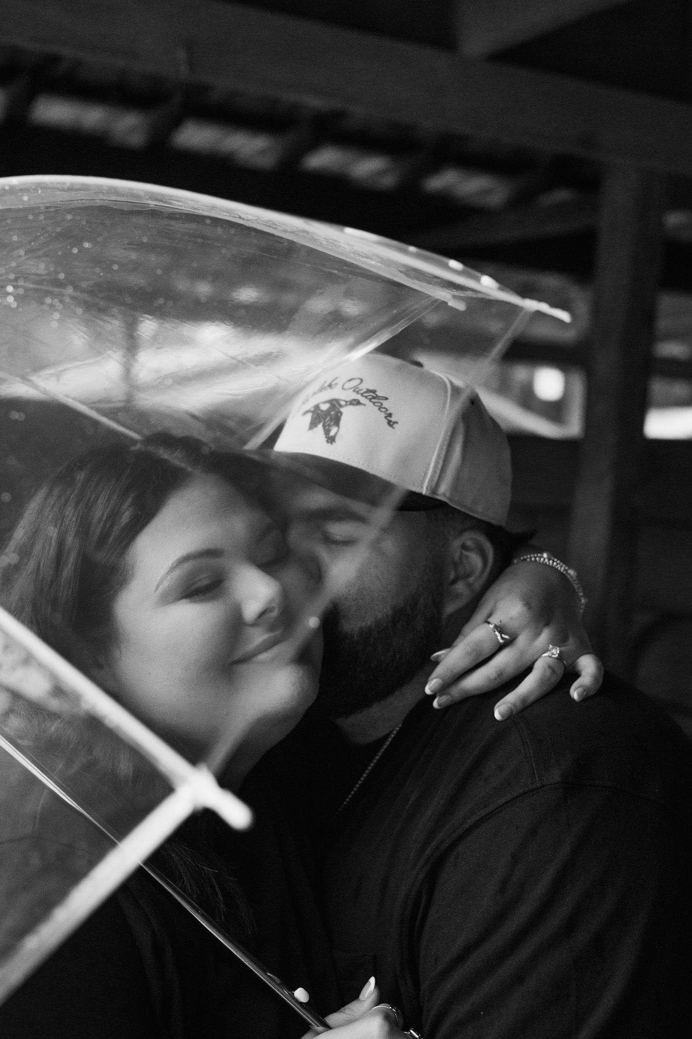 Black and white photo of a couple sharing a kiss under a clear umbrella, woman wearing a ring embracing a bearded man.