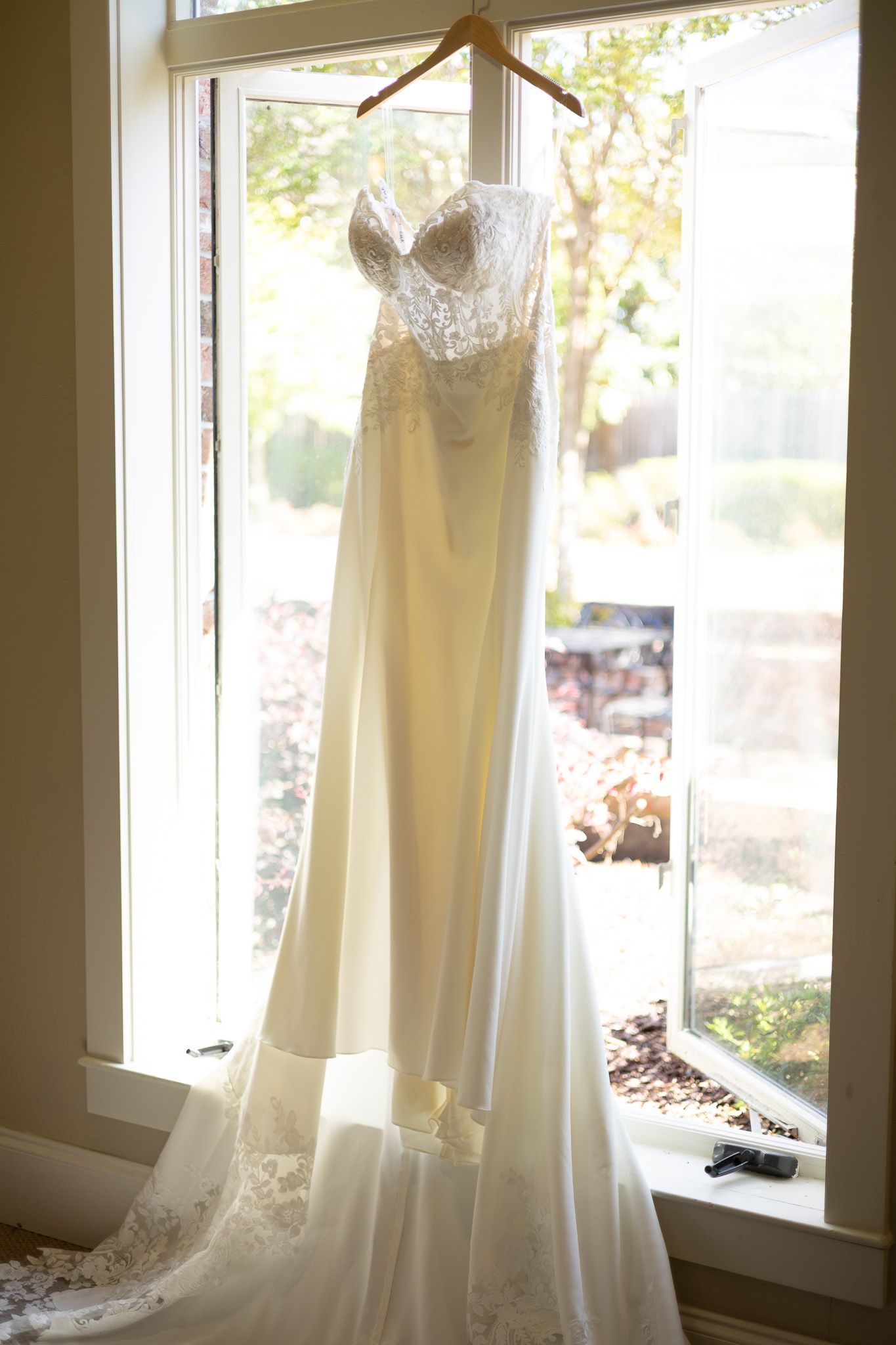 Elegant strapless lace wedding gown hanging on a wooden hanger in front of a sunlit window.