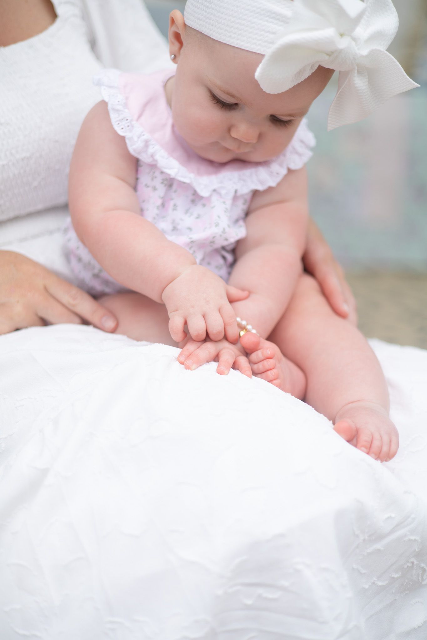 Adorable baby in pink floral outfit with white bow examines her tiny feet and bracelet.