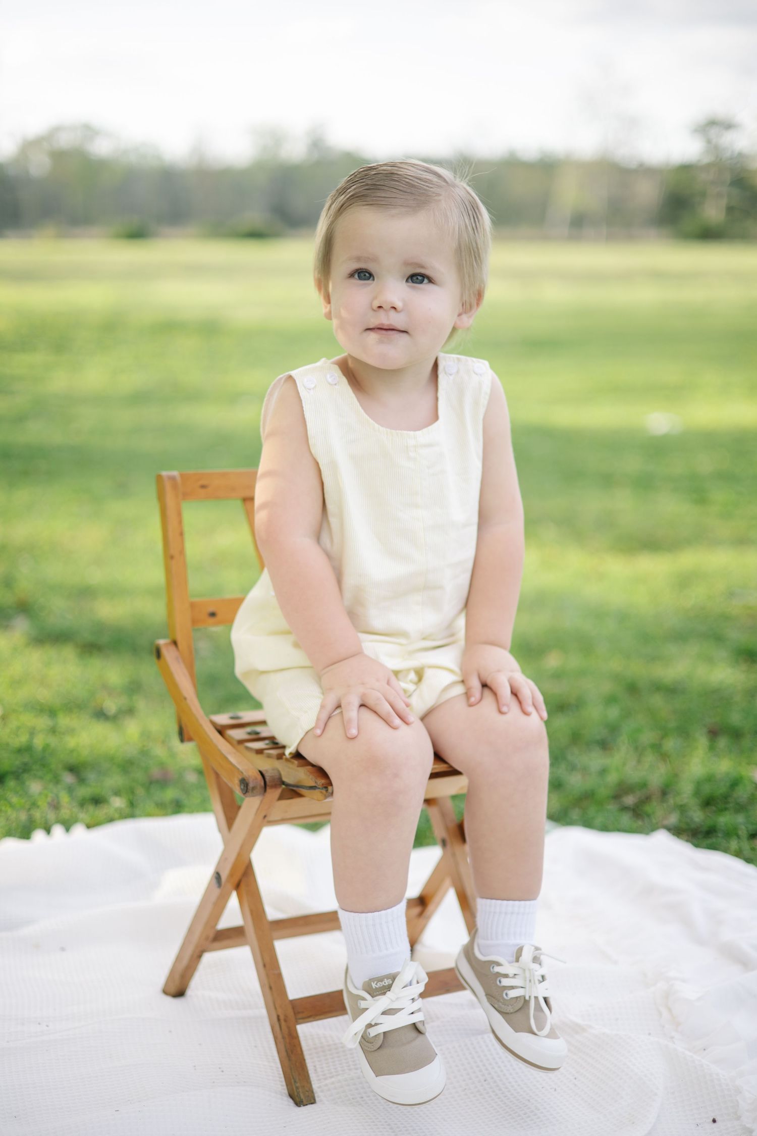 Cute toddler in a yellow romper sitting on a wooden folding chair in a green field.