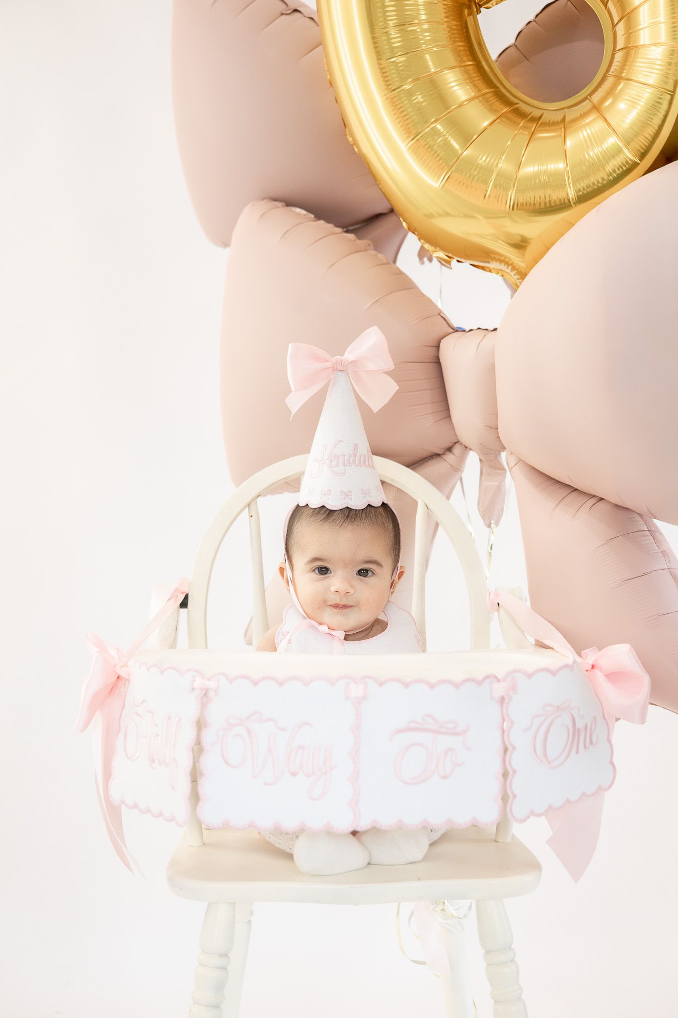 Baby girl in birthday hat sits in white chair with pink balloons and gold number one balloon for first birthday.
