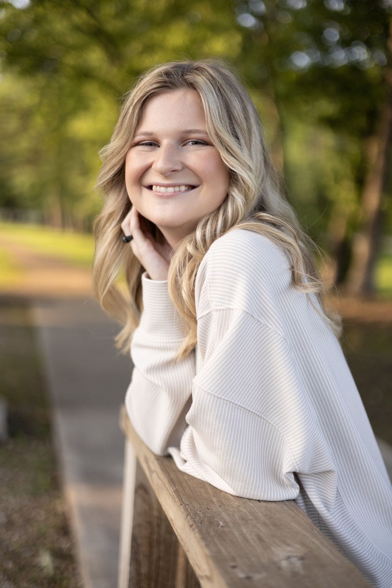 Happy blonde teenage girl in a cream sweater smiles while leaning on a wooden fence outdoors.