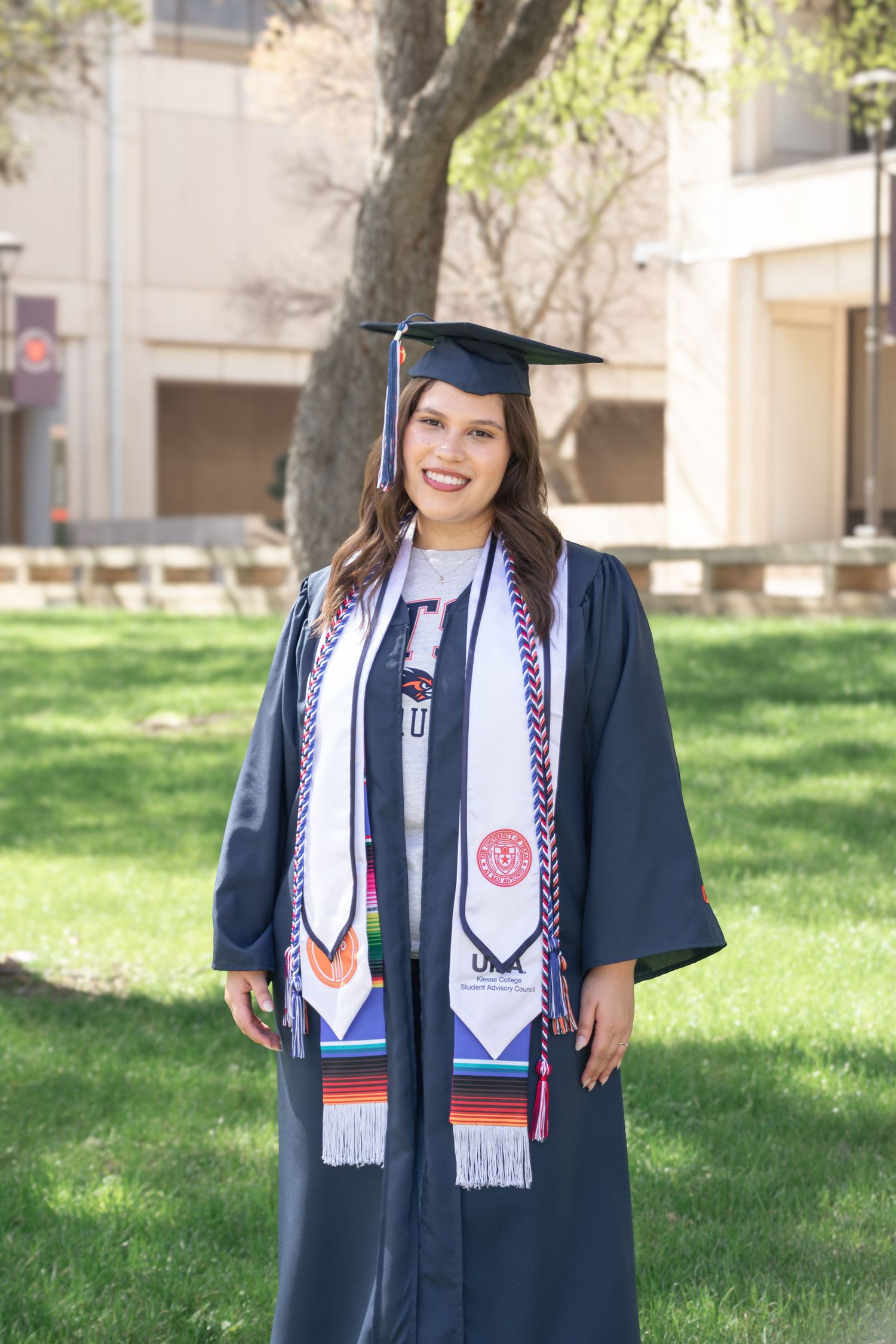 UTSA graduation portrait on campus lawn in cap gown and honor stoles in San Antonio