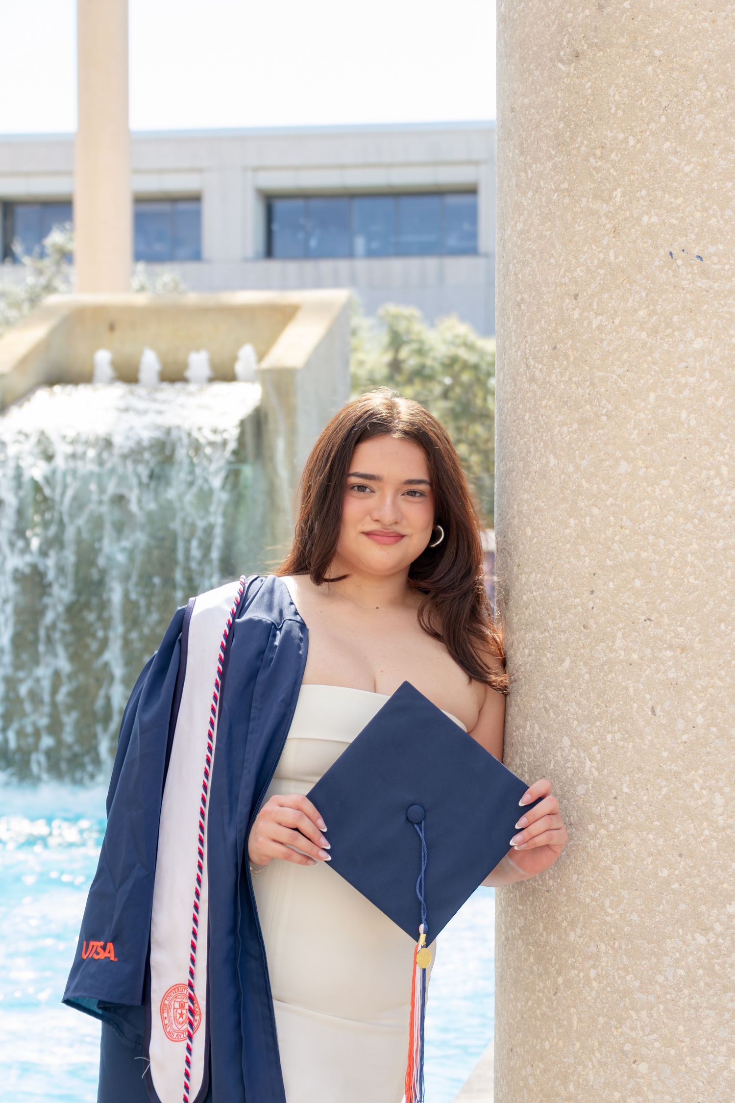 UTSA graduation portrait at the Sombrilla fountain holding cap and gown with honor stoles