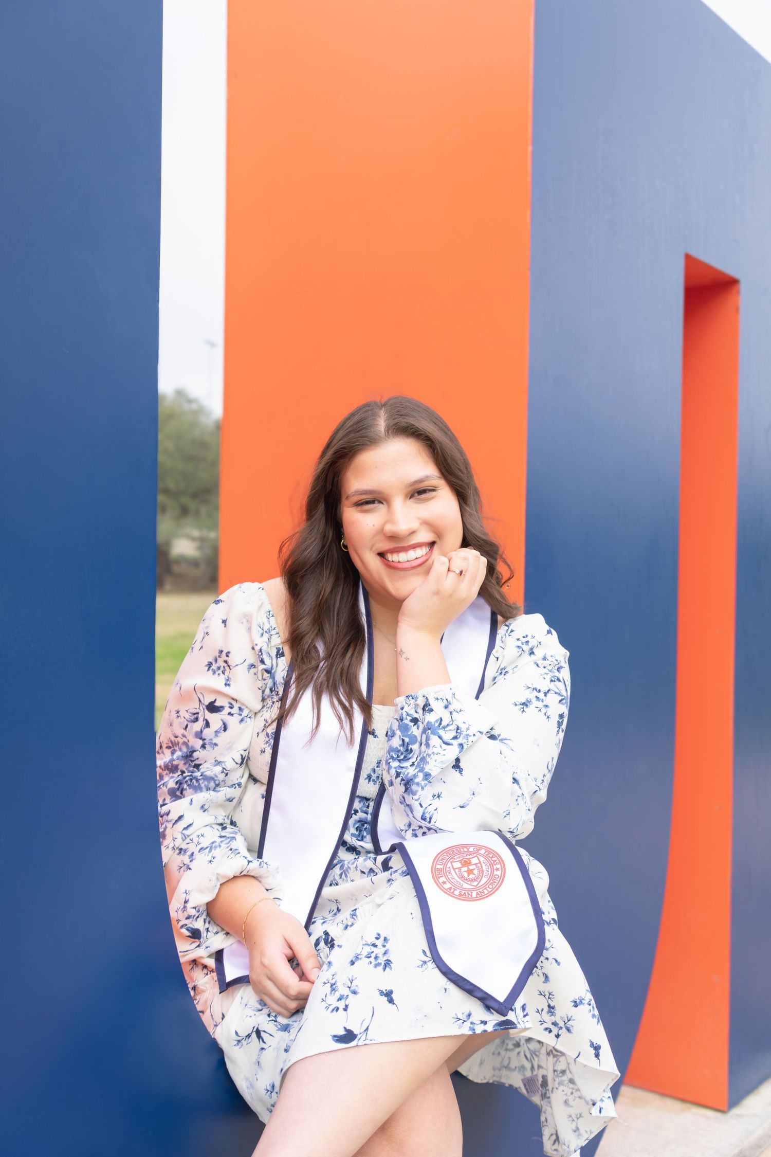 UTSA graduation portrait leaning against the UTSA letter sculptures in San Antonio in floral dress