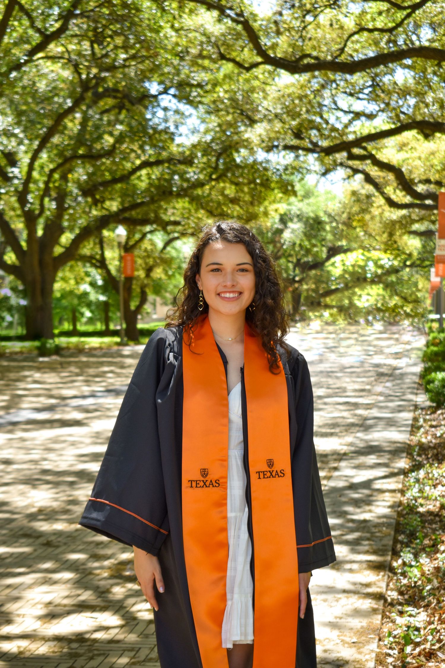 UT Austin graduation portrait along tree-lined campus walk in cap gown and orange stole