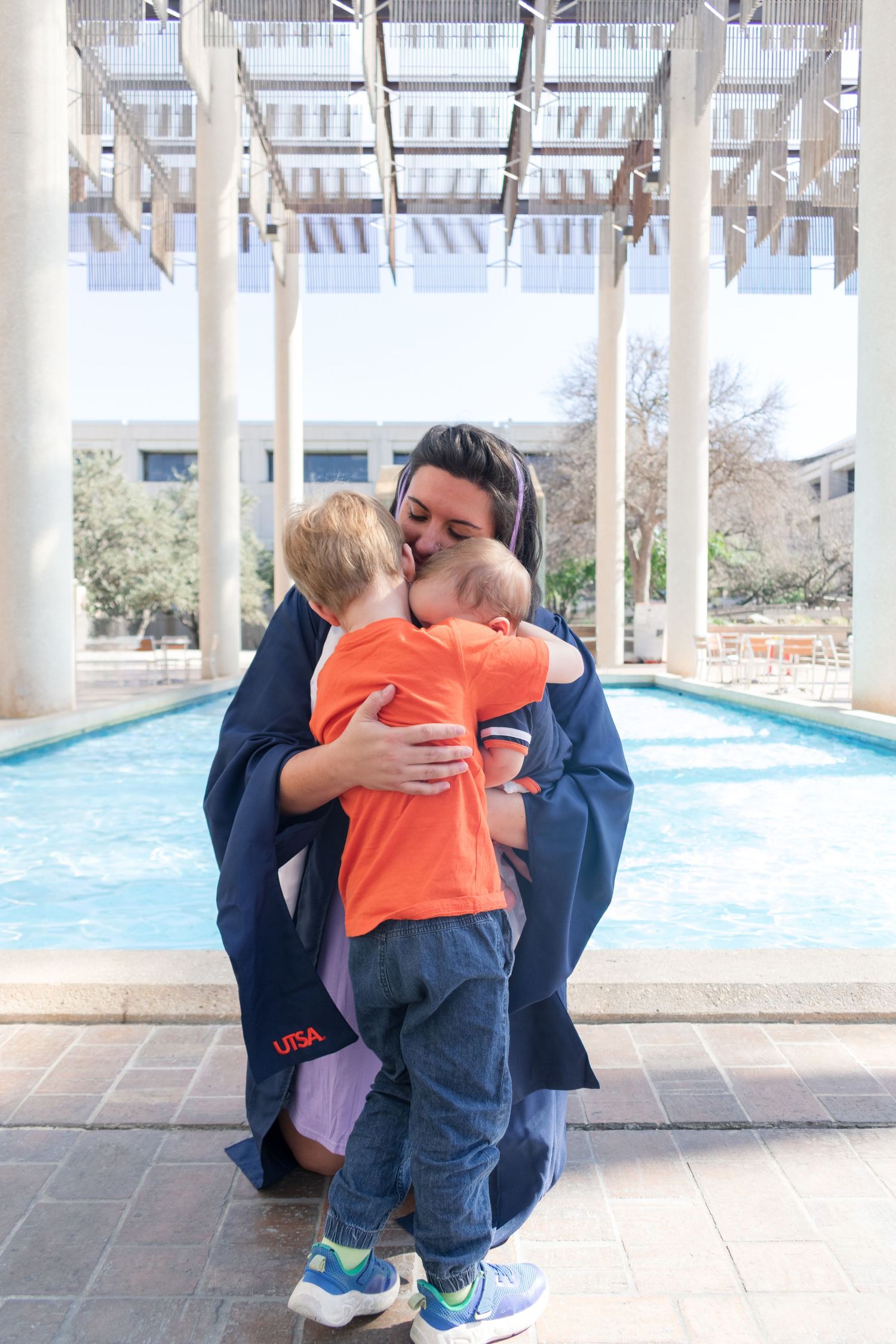 UTSA graduation portrait at the Sombrilla fountain with graduate hugging young child in cap and gown