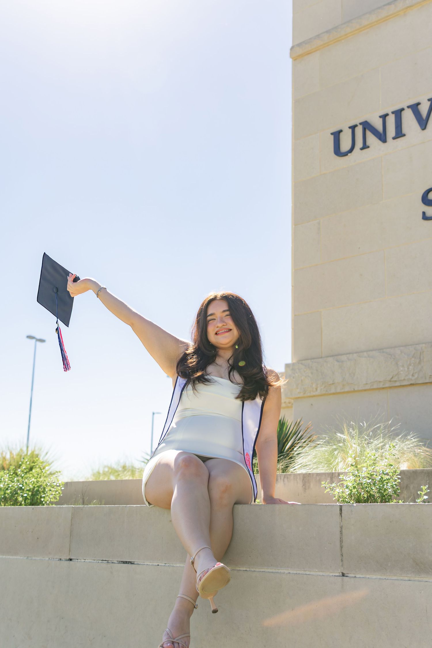 UTSA graduation portrait seated at the University of Texas San Antonio sign holding cap