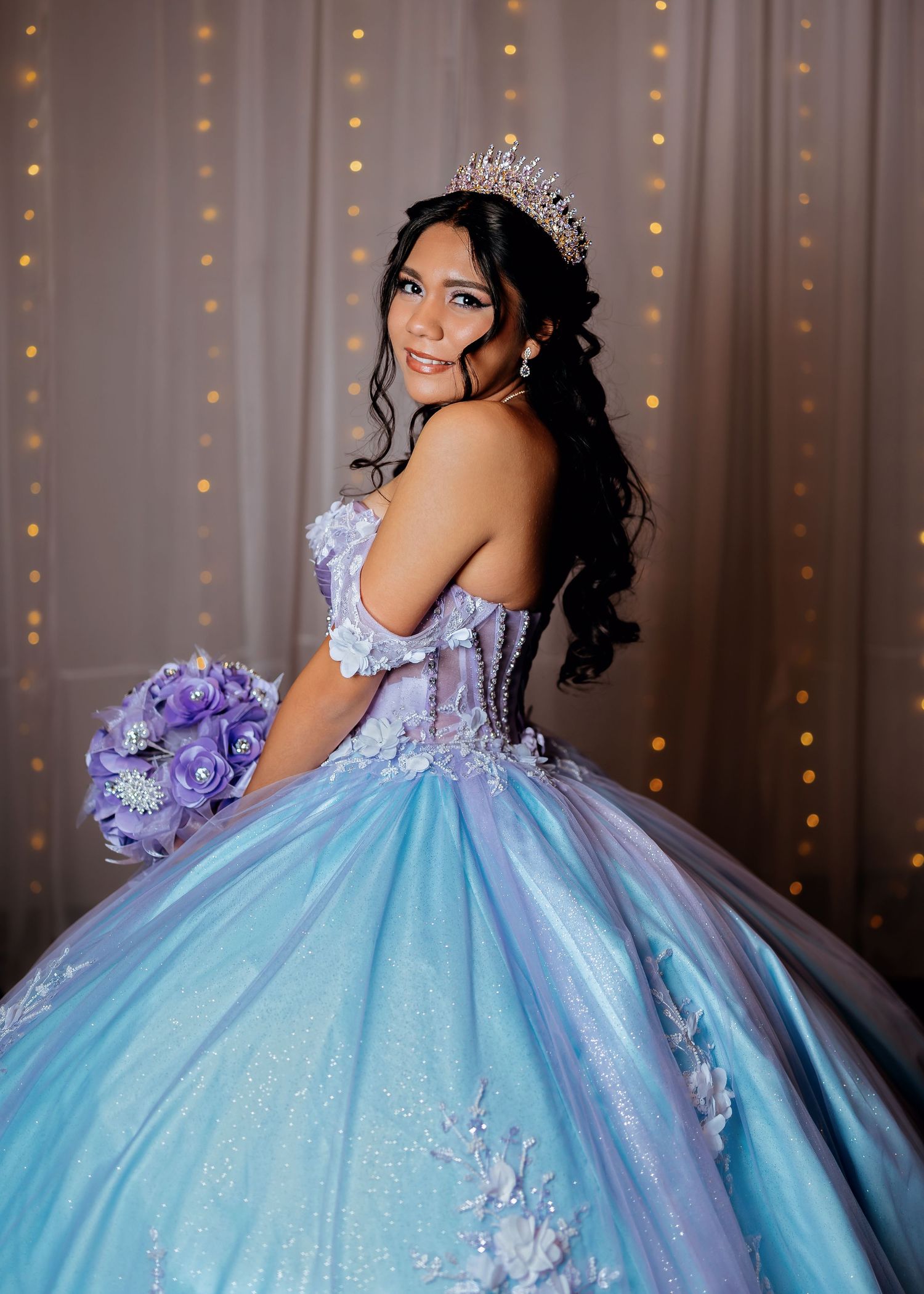 Beautiful quinceañera in blue and lavender ball gown with tiara holding purple floral bouquet.