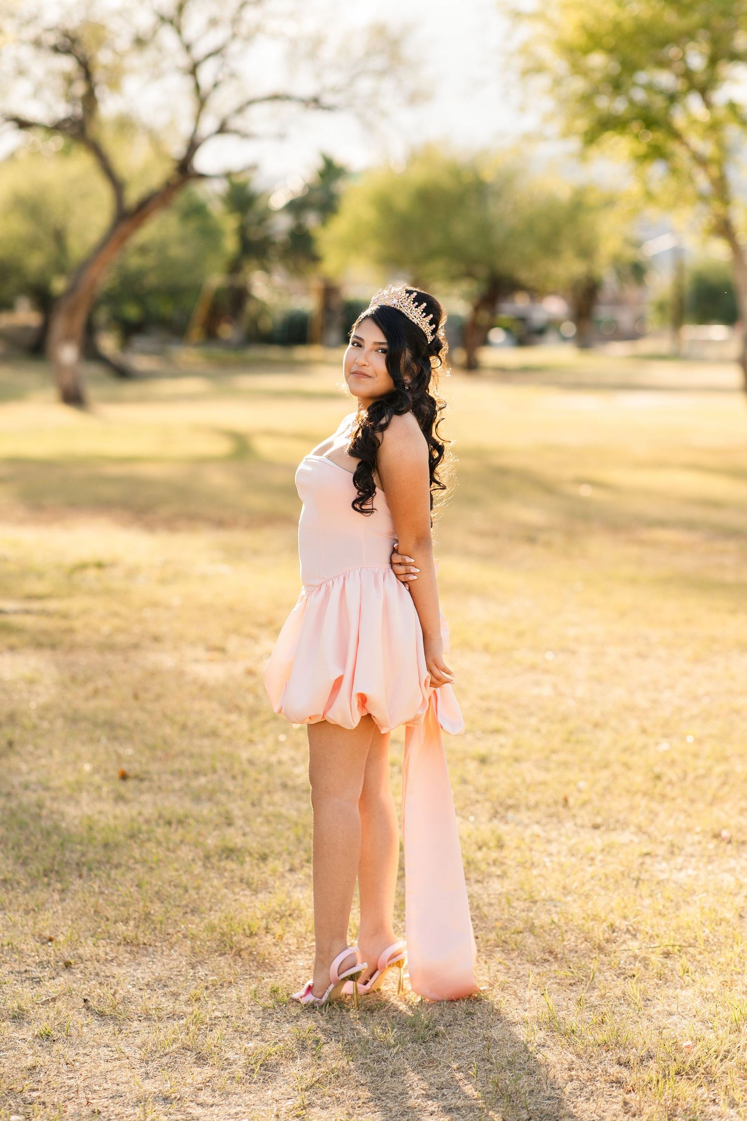 Teen girl in blush pink quinceanera dress with train poses in sunlit park during portrait session.