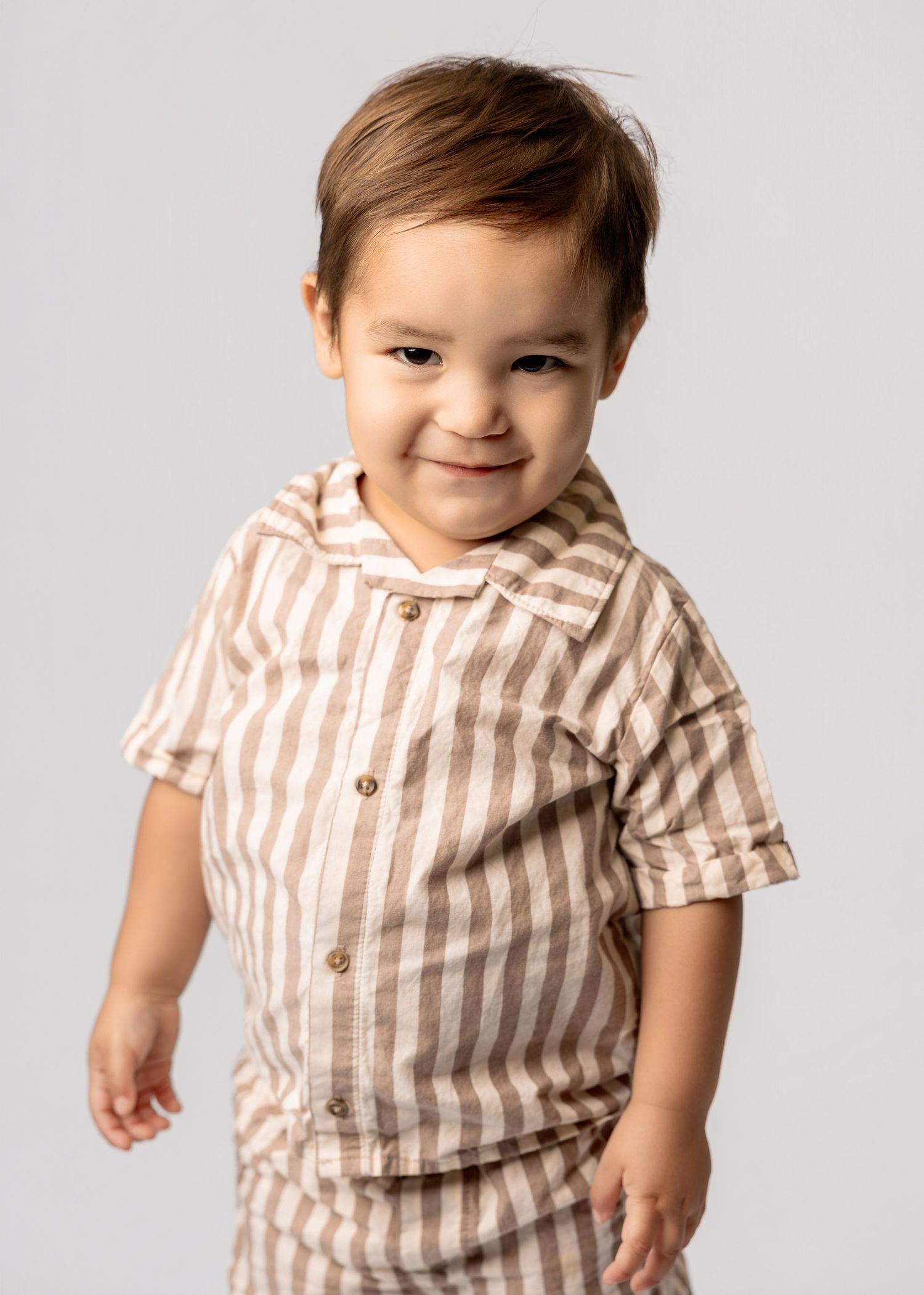 Toddler boy smiling in a beige and white striped short-sleeve button-up shirt on a gray background.