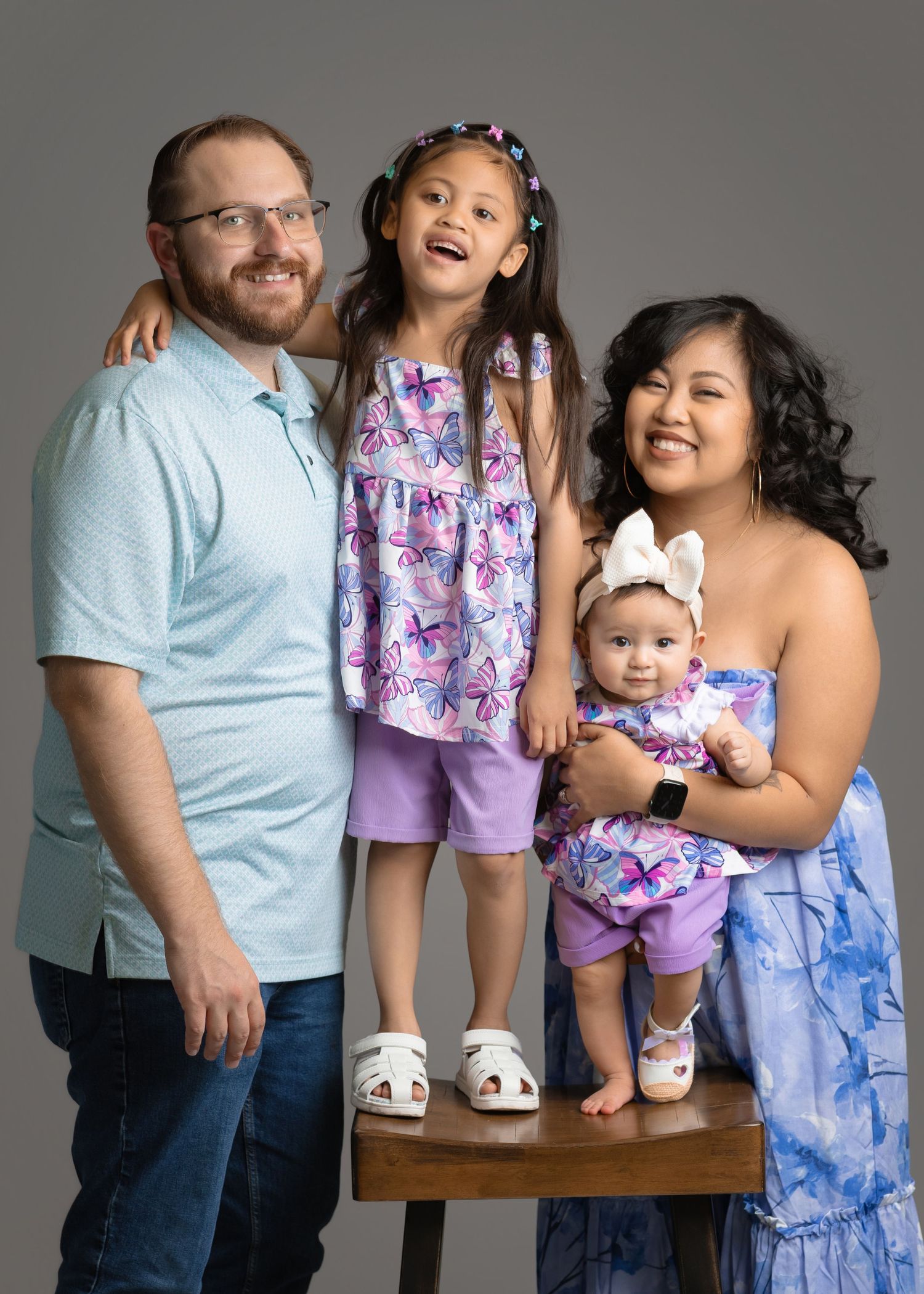 Happy family of four posing in studio with two young daughters in purple floral outfits.