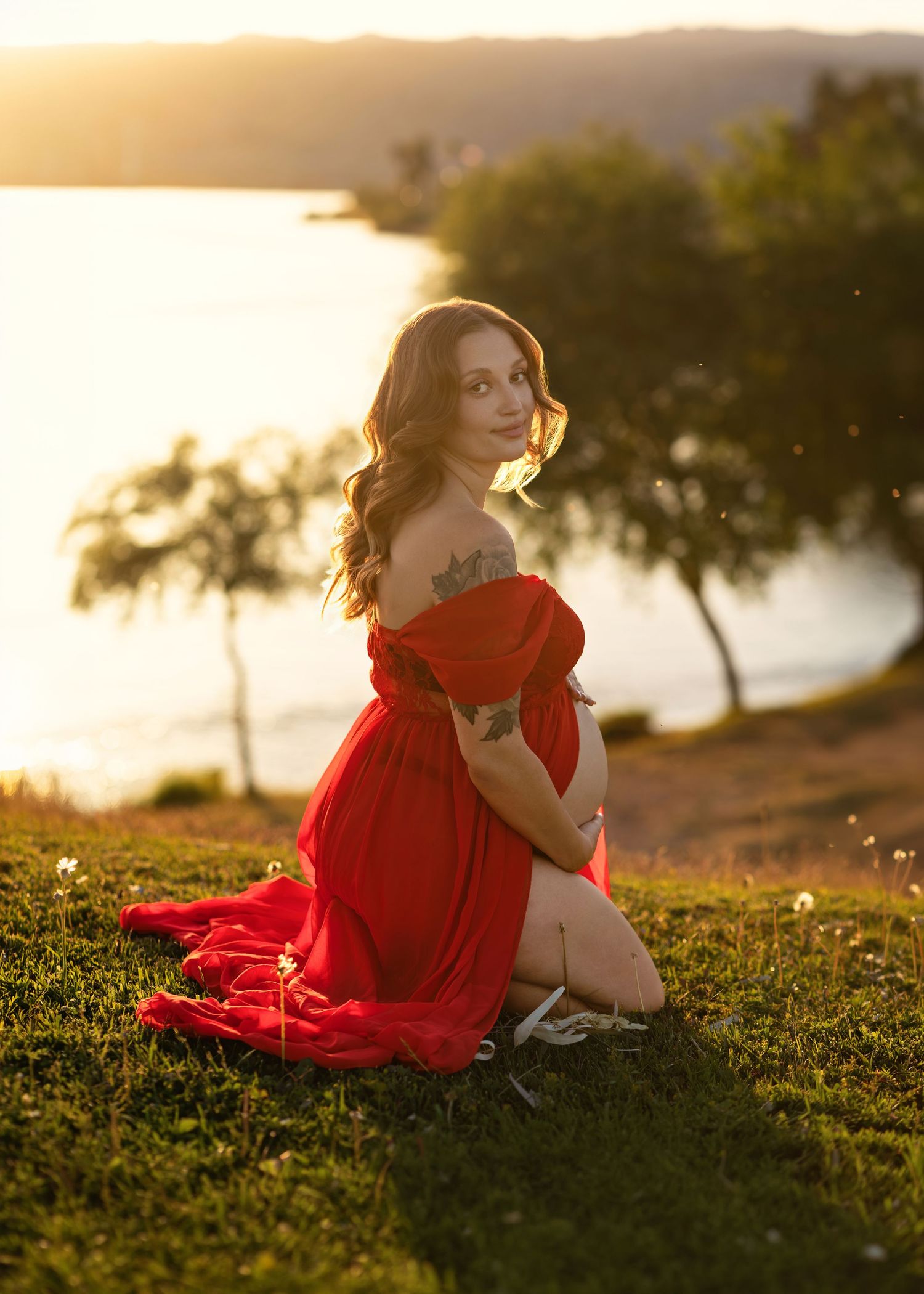 Pregnant woman in red off-shoulder gown sitting on grass at golden hour near a lake.