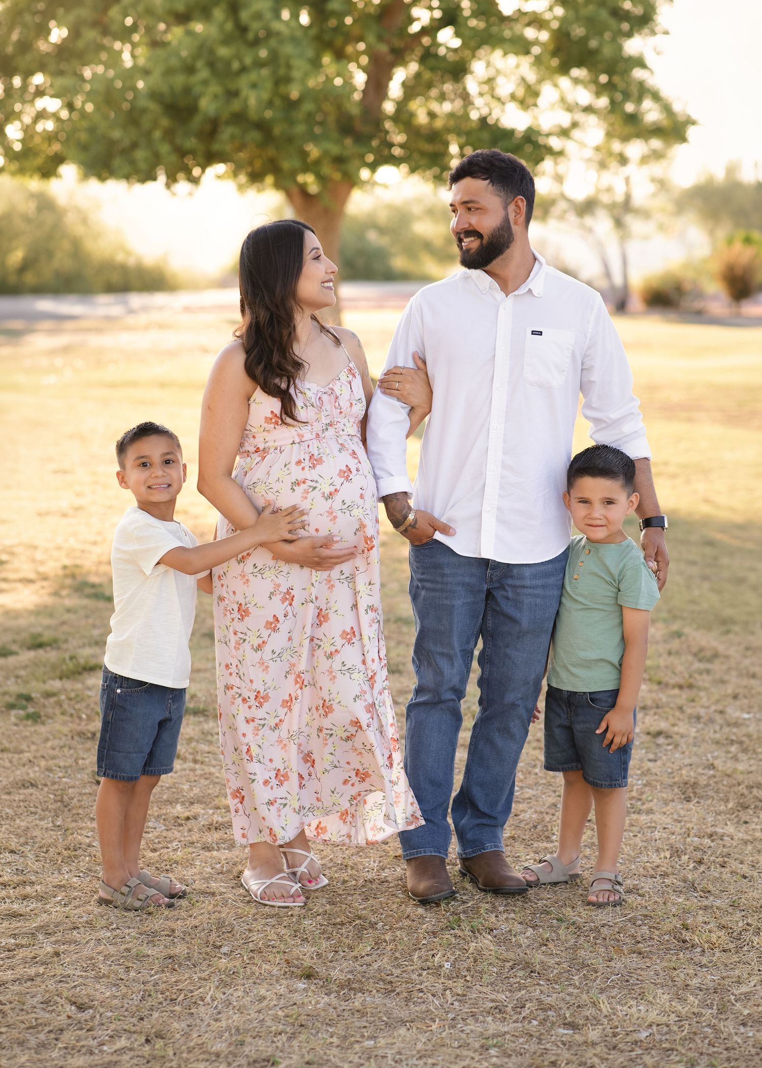 Family maternity photo with pregnant mom, dad, and two young sons in a sunny park.