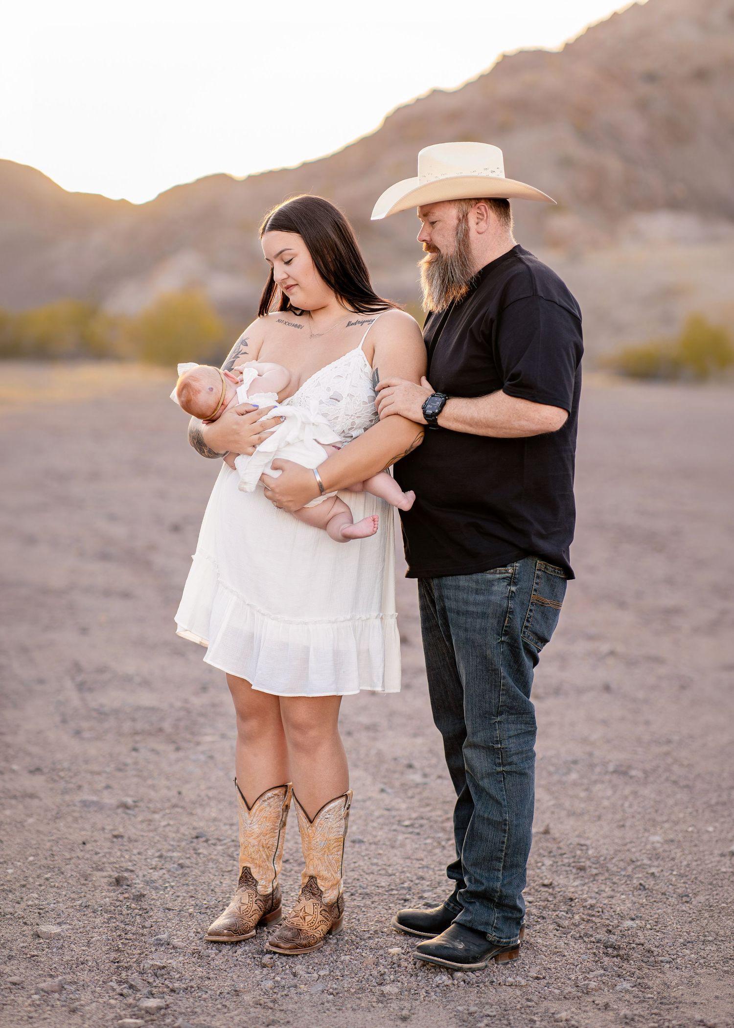New parents in desert landscape tenderly gaze at newborn baby at golden hour, woman wearing cowboy boots.