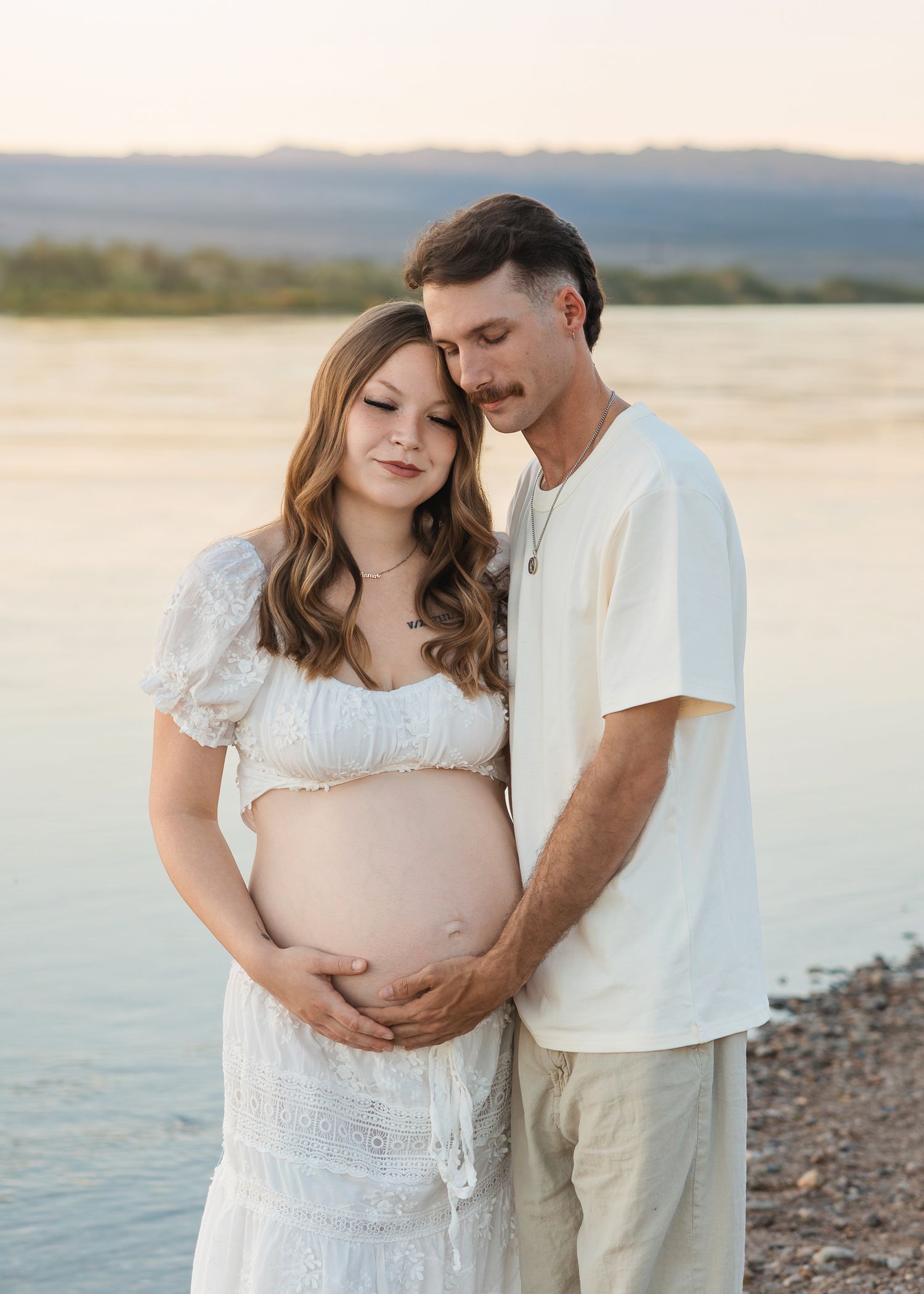 Pregnant couple in white outfits embracing by a lake at golden hour, cradling baby bump during maternity photo session.