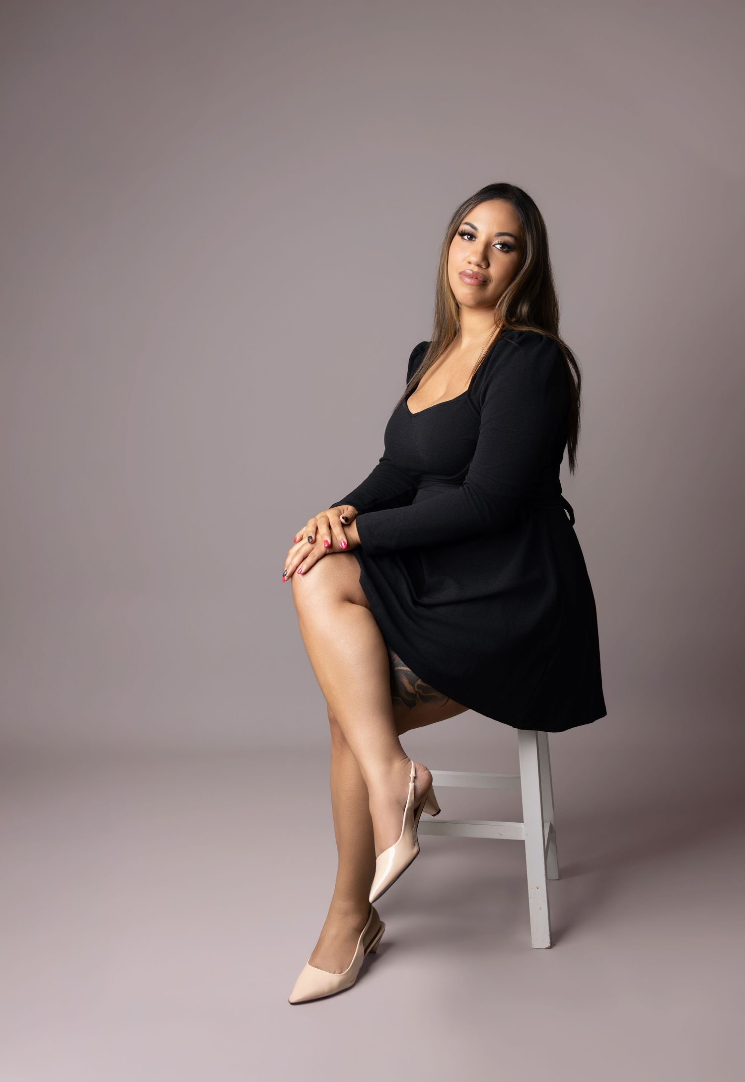 A woman in a black dress and nude heels sits elegantly on a white stool against a gray background.