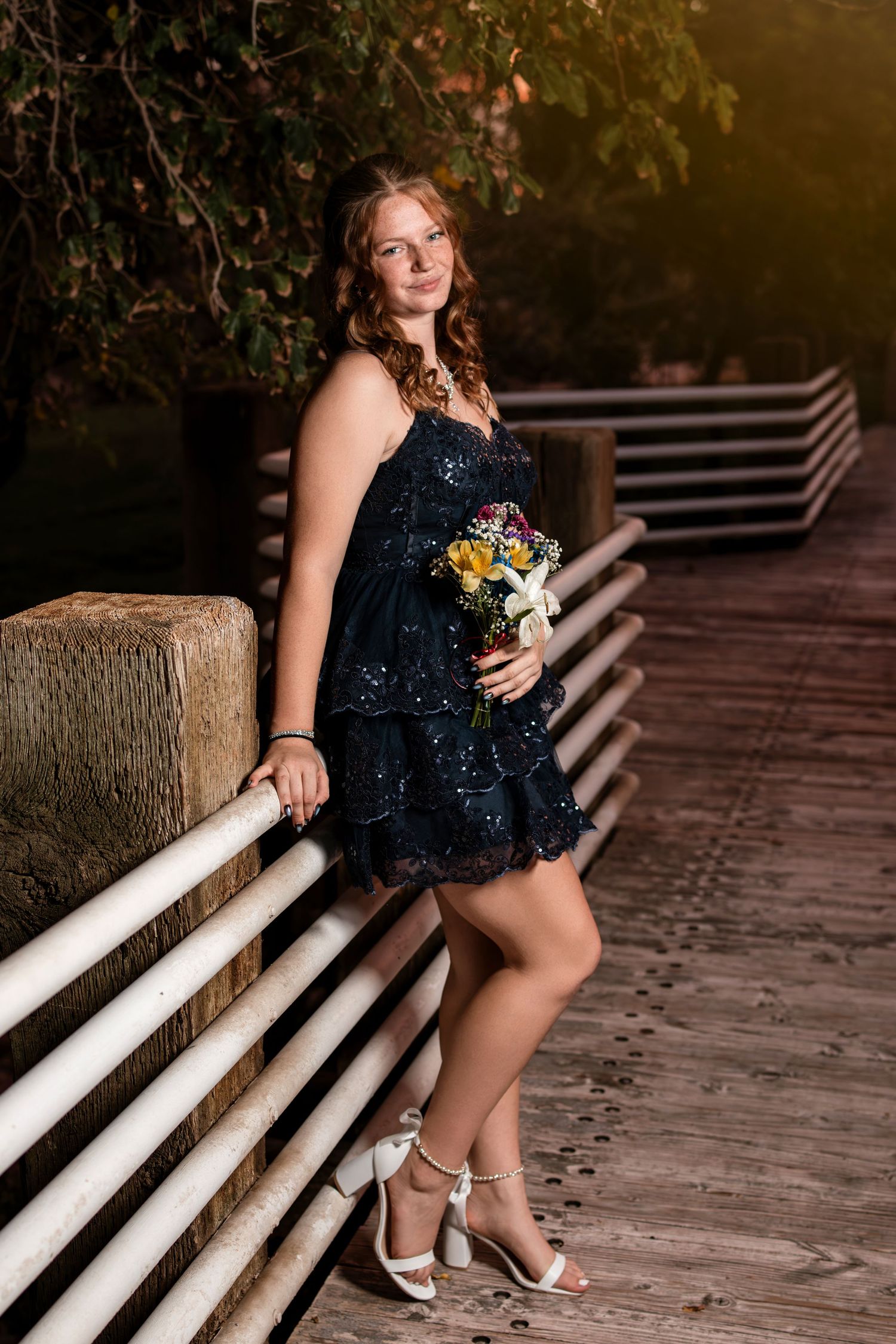 Young woman in navy sequin dress holds floral bouquet on wooden bridge at night.