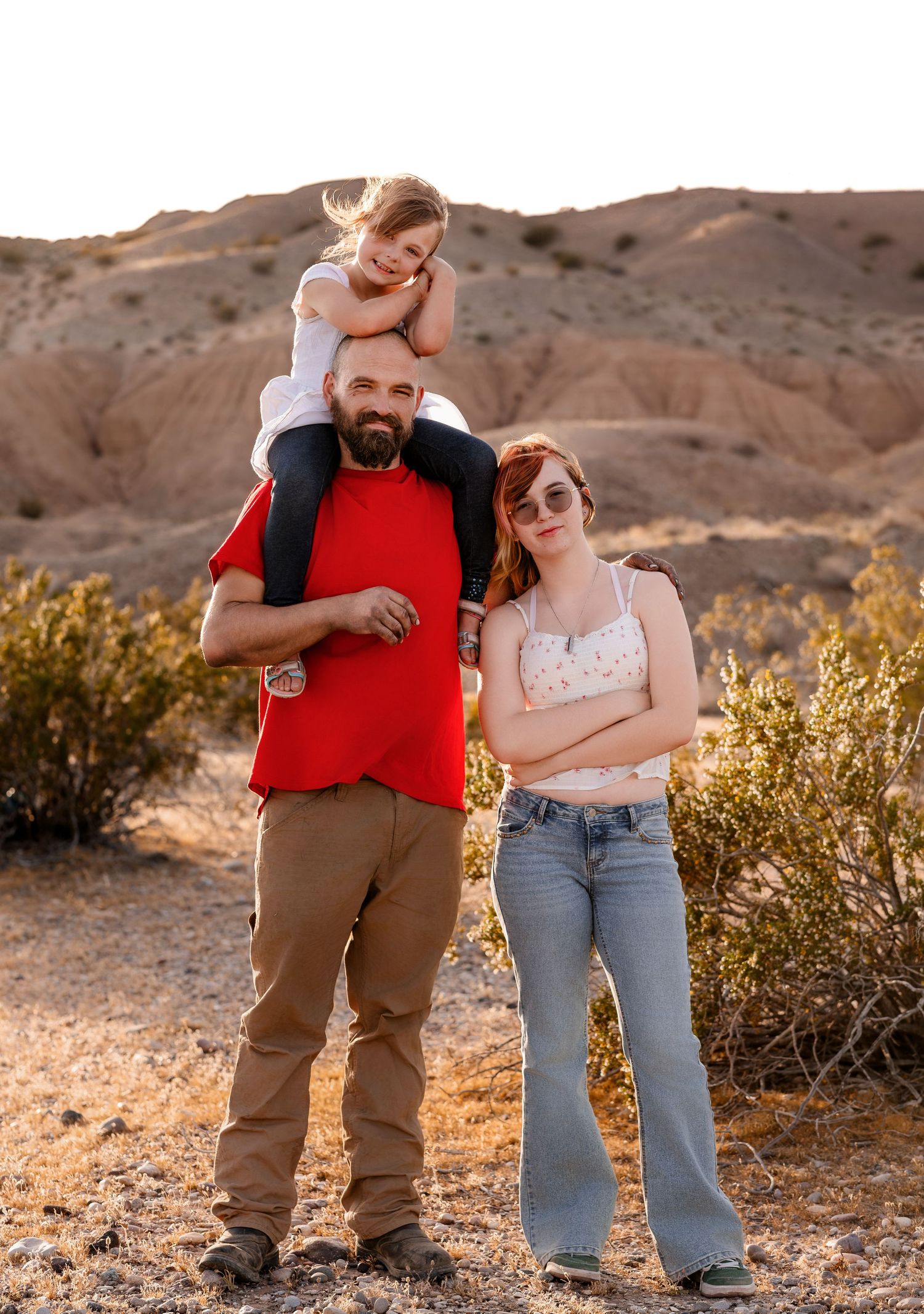 A bald bearded man in a red shirt carries a young girl on his shoulders beside a teenage girl outdoors.