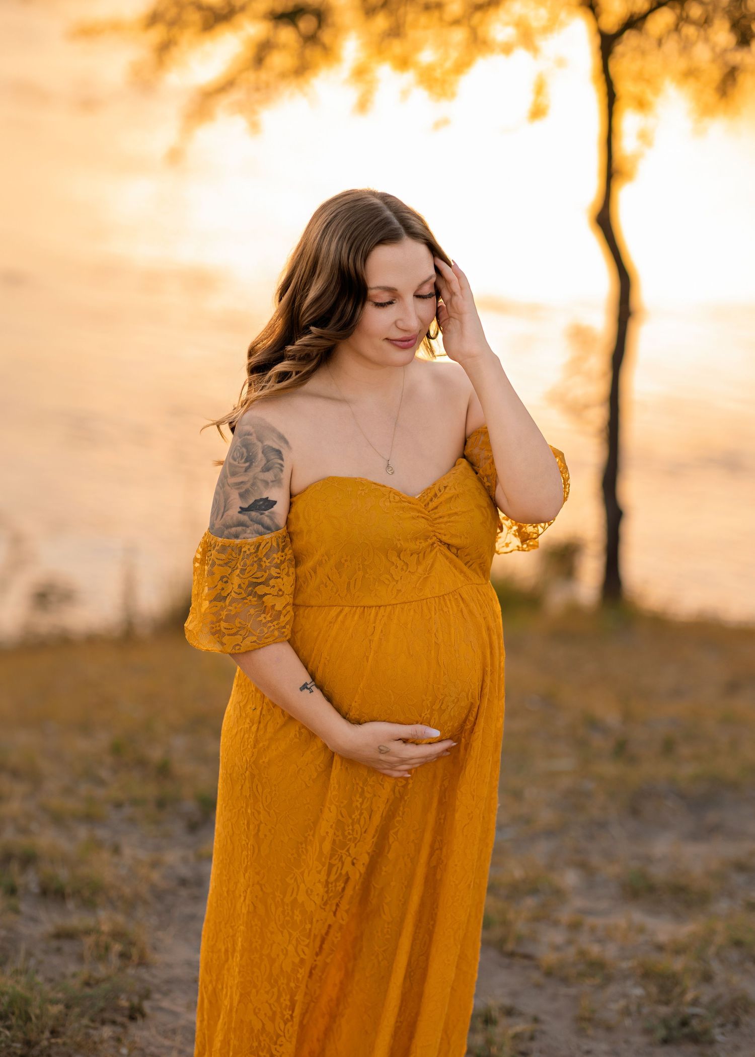 A pregnant woman in a mustard yellow off-shoulder lace gown poses outdoors at golden hour.