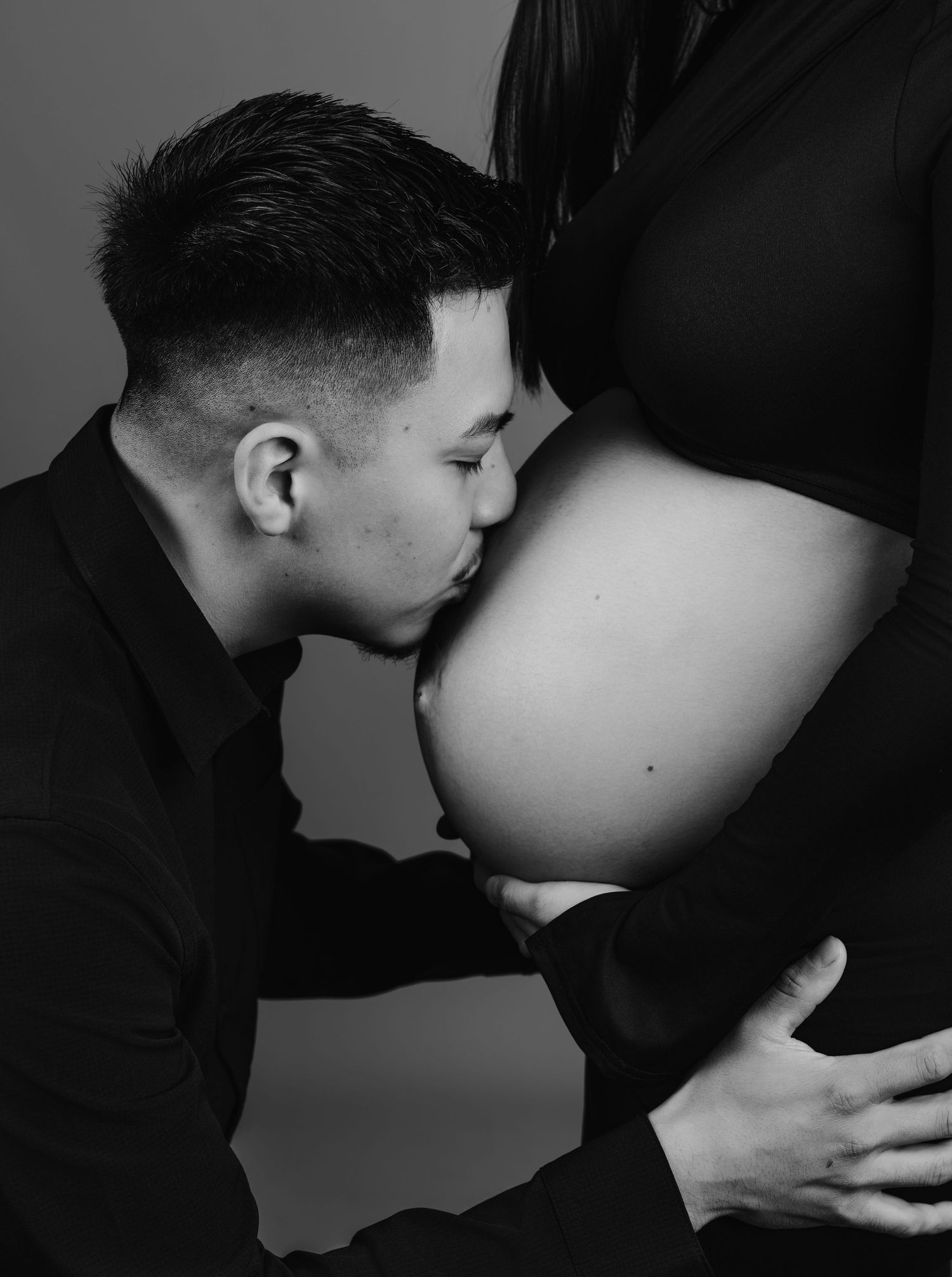 Black and white photo of a man kissing pregnant woman's baby bump in a studio setting.