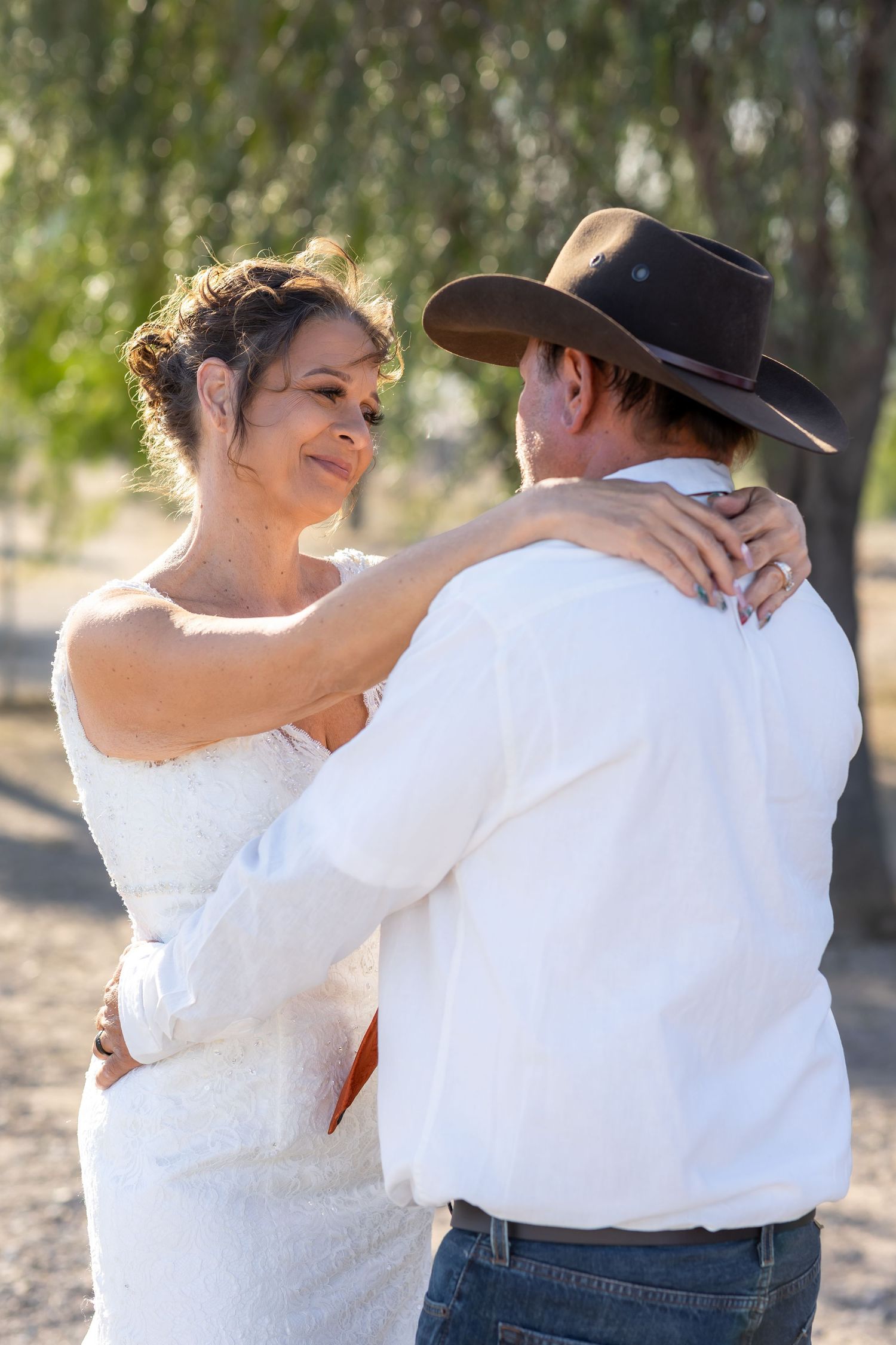 Bride in lace wedding dress embracing groom in cowboy hat outdoors under trees.
