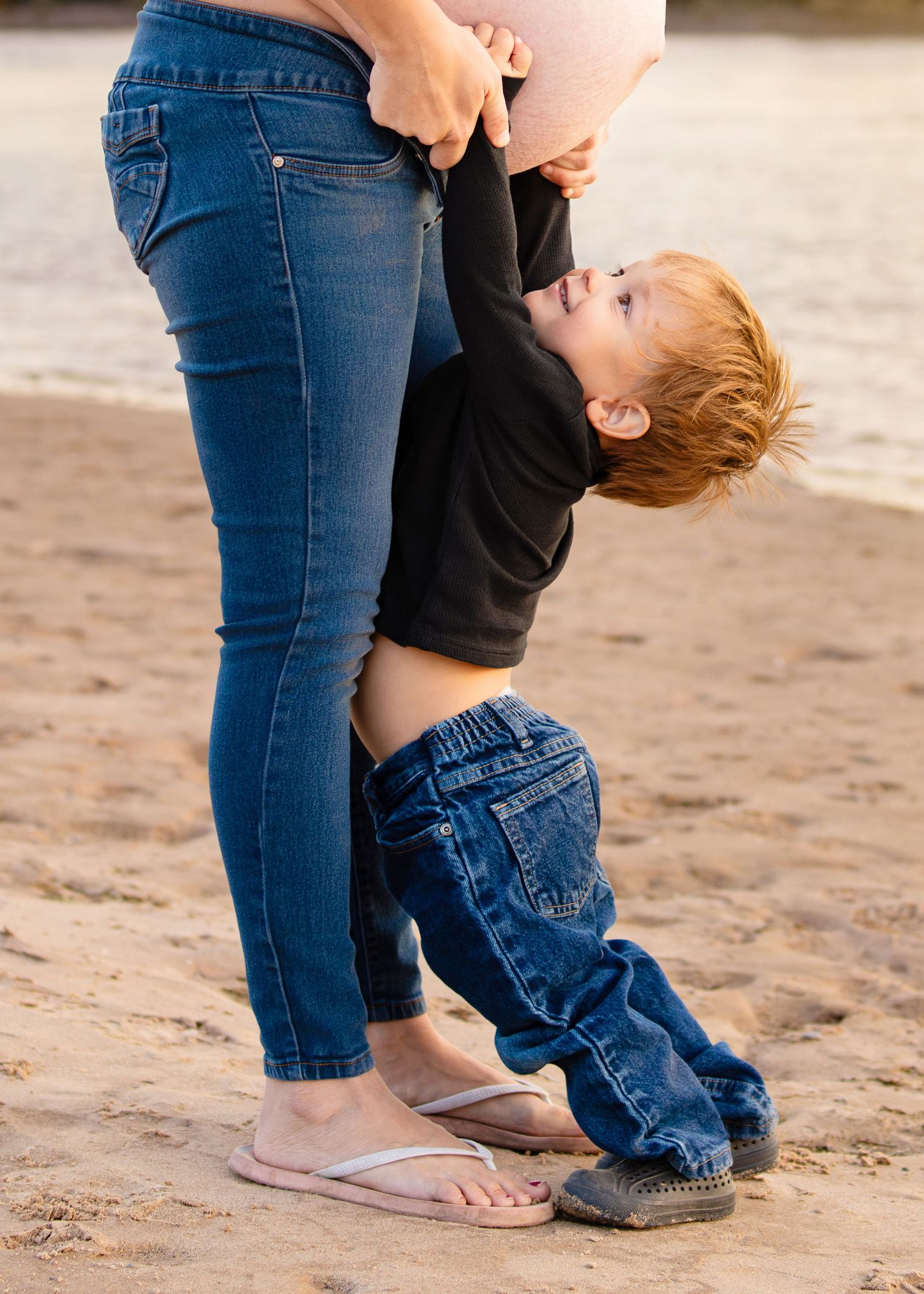 Young redhead boy playfully hugging pregnant mothers baby bump on sandy beach.