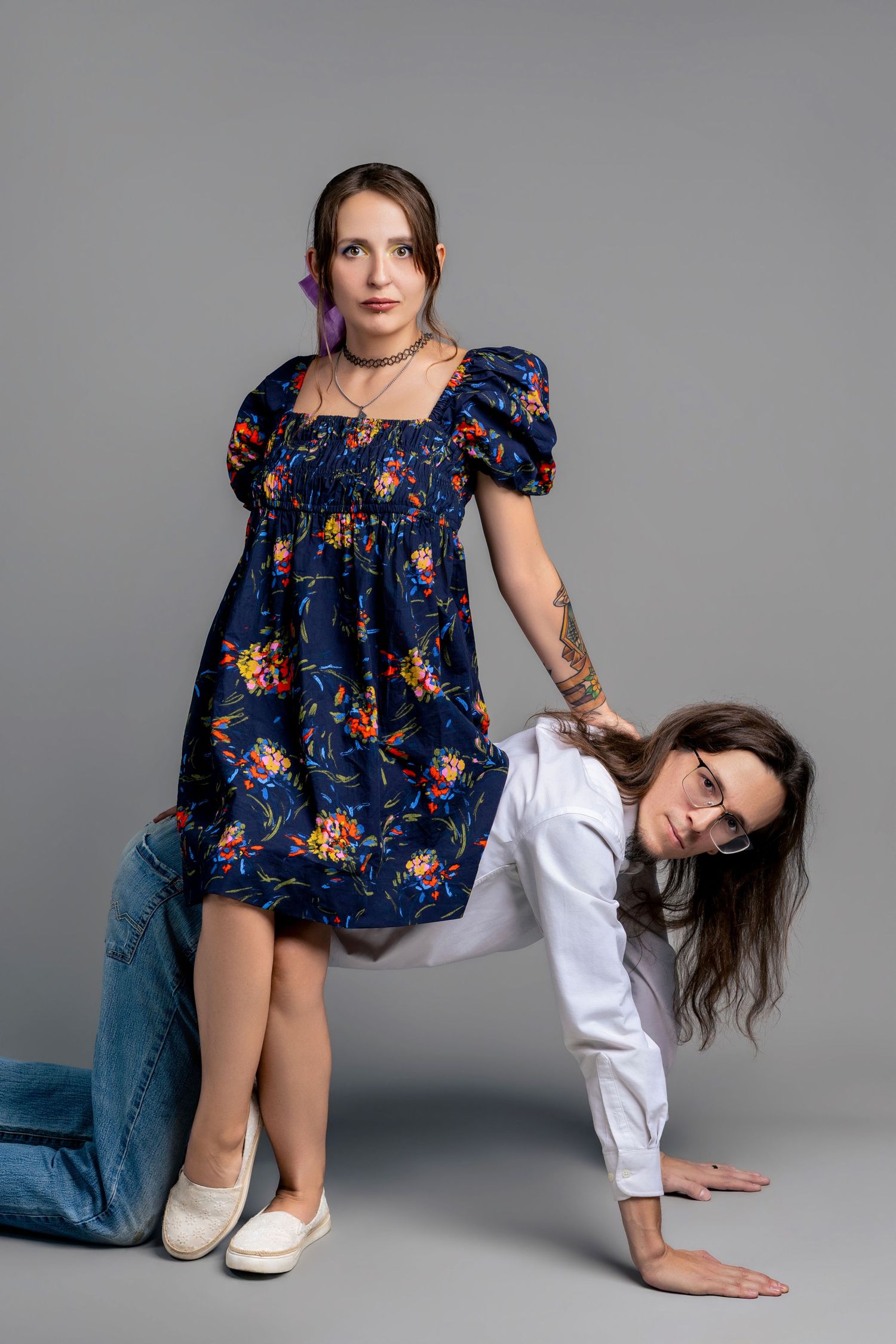 Two women pose in a studio, one in a floral blue dress standing while the other in white crouches beside her.