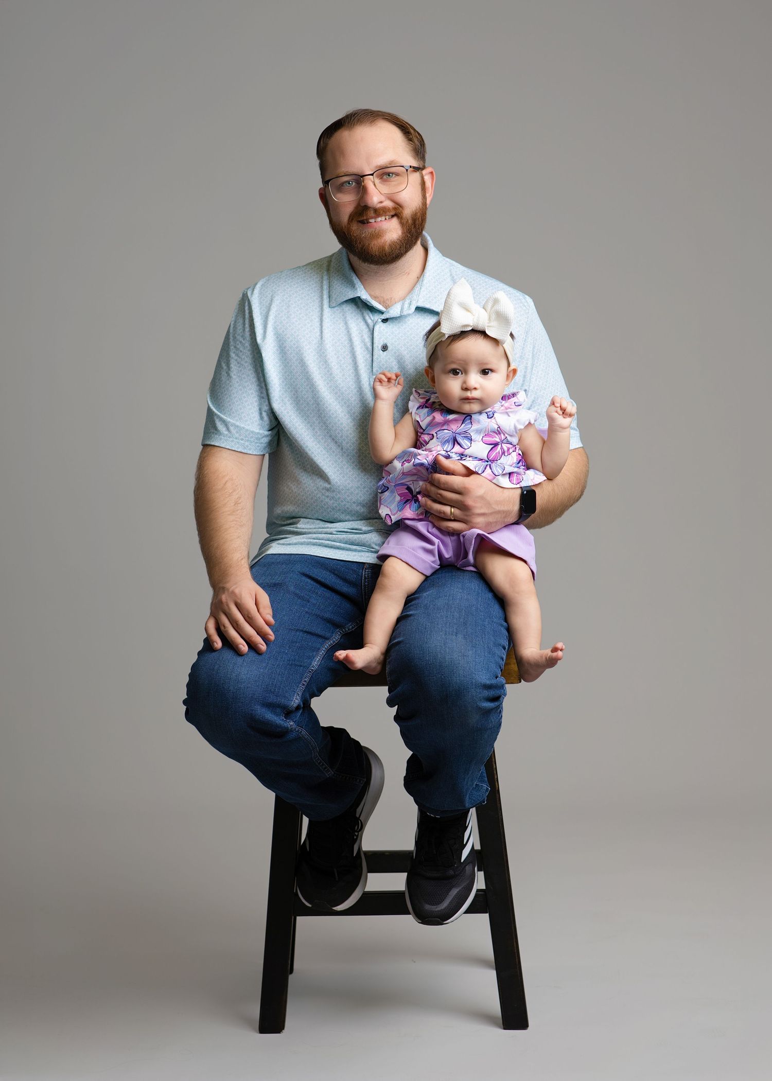 Smiling bearded man in glasses and a light blue polo sits on a black stool holding a baby girl in a purple floral outfit and white bow.