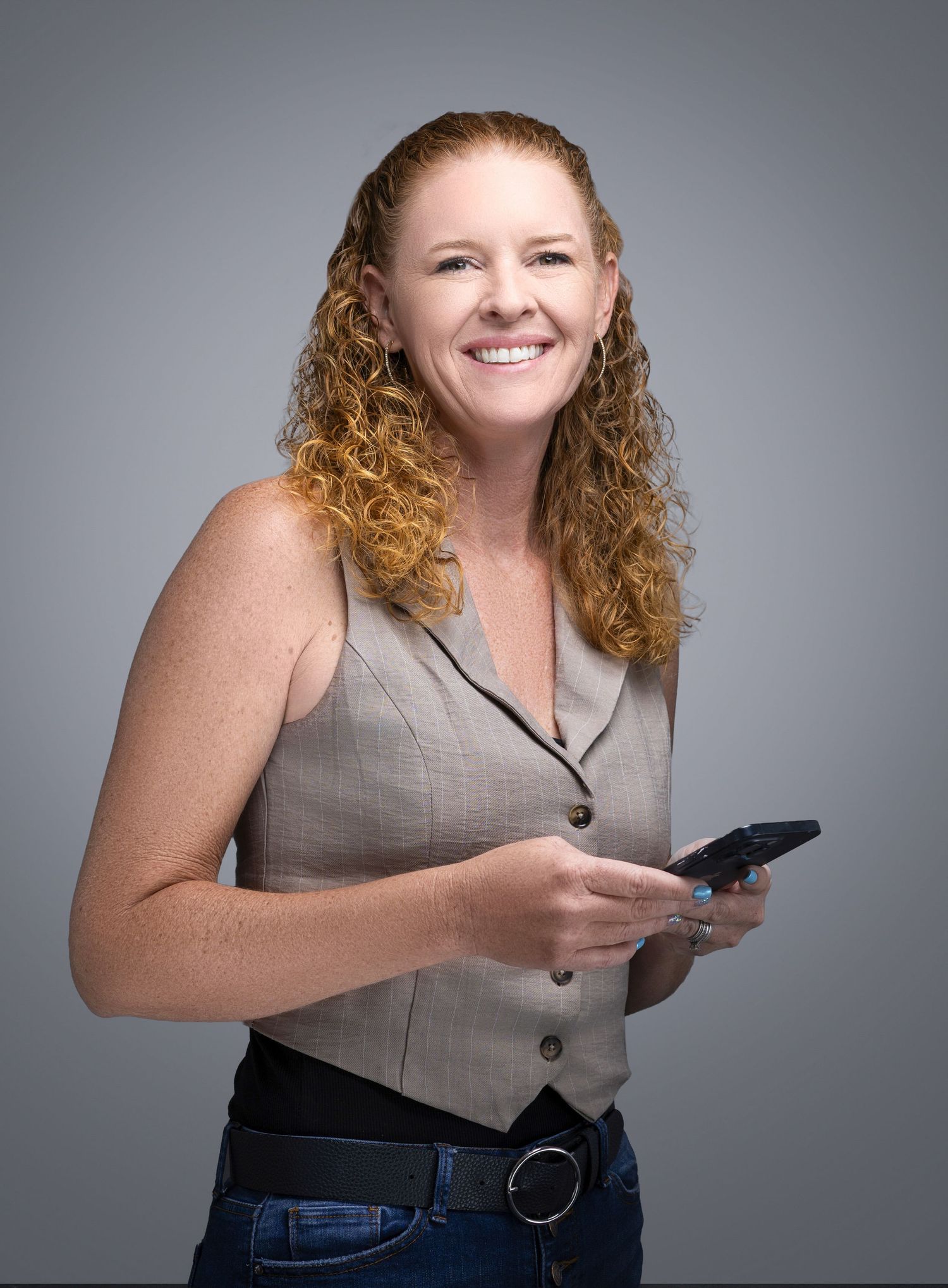 Professional woman with curly red hair smiling while holding smartphone in gray vest.