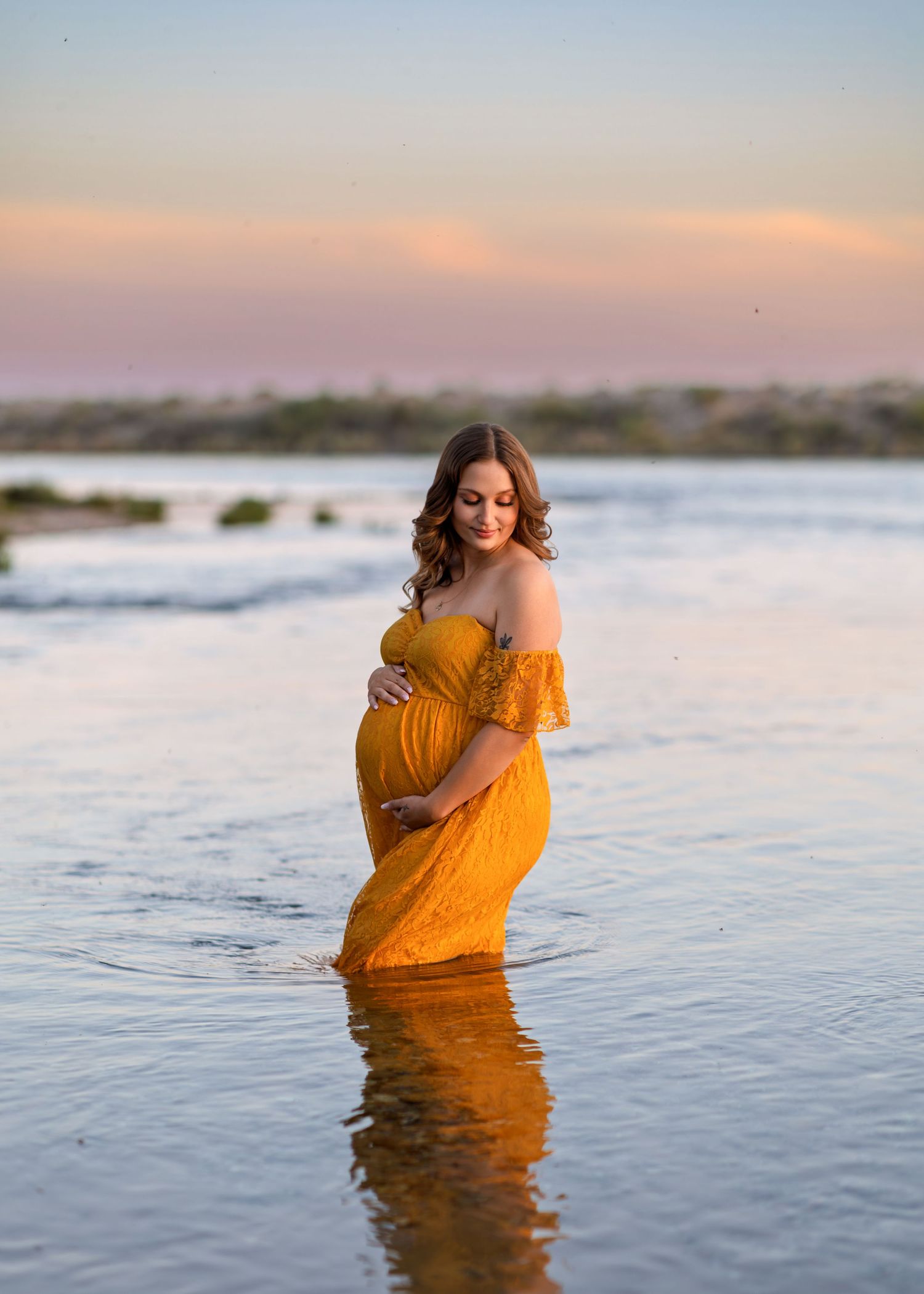 A pregnant woman in a golden yellow lace gown stands in shallow water at sunset.