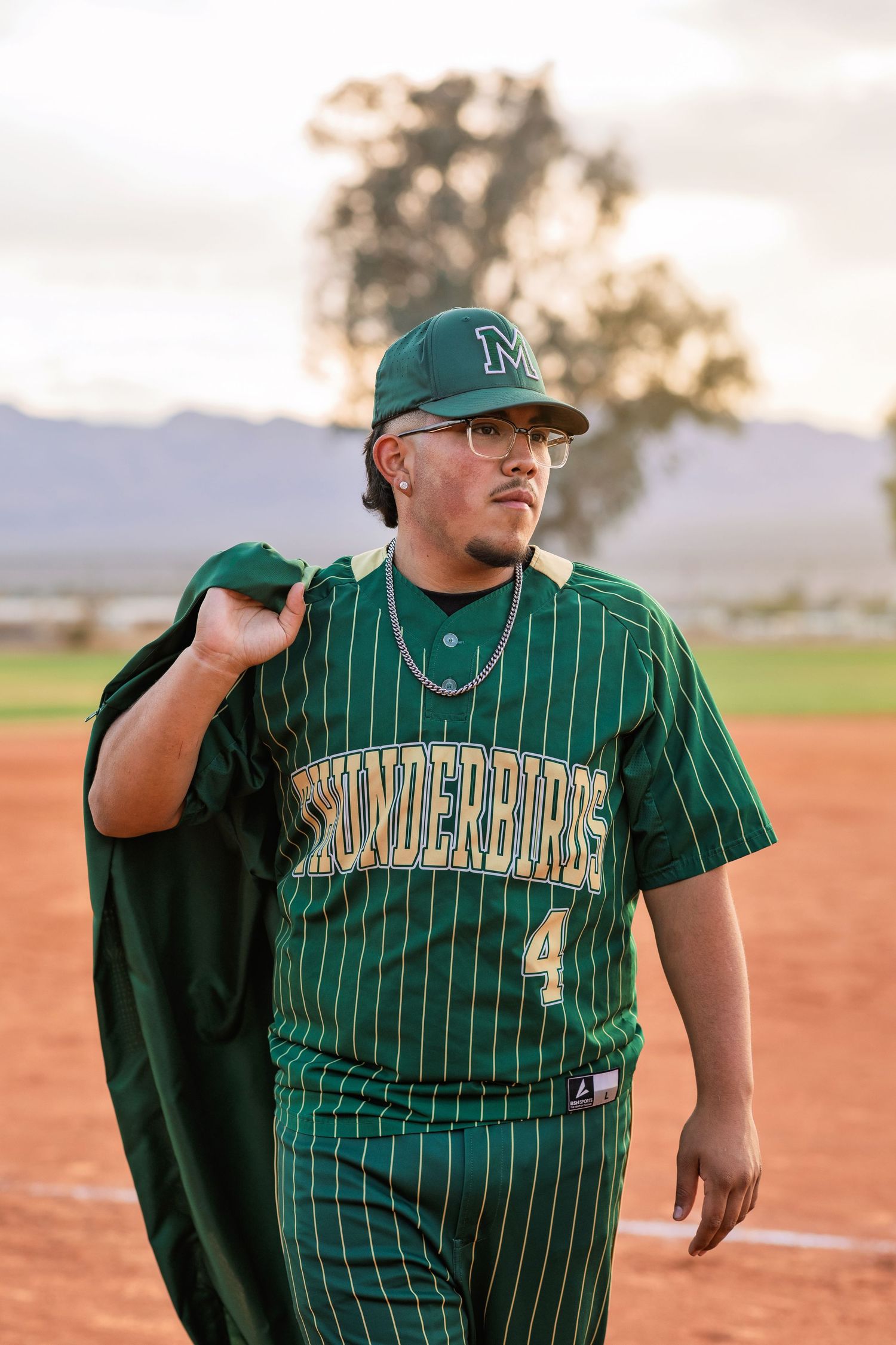 Young baseball player in green Thunderbirds pinstripe uniform number 4 walking on field at sunset.