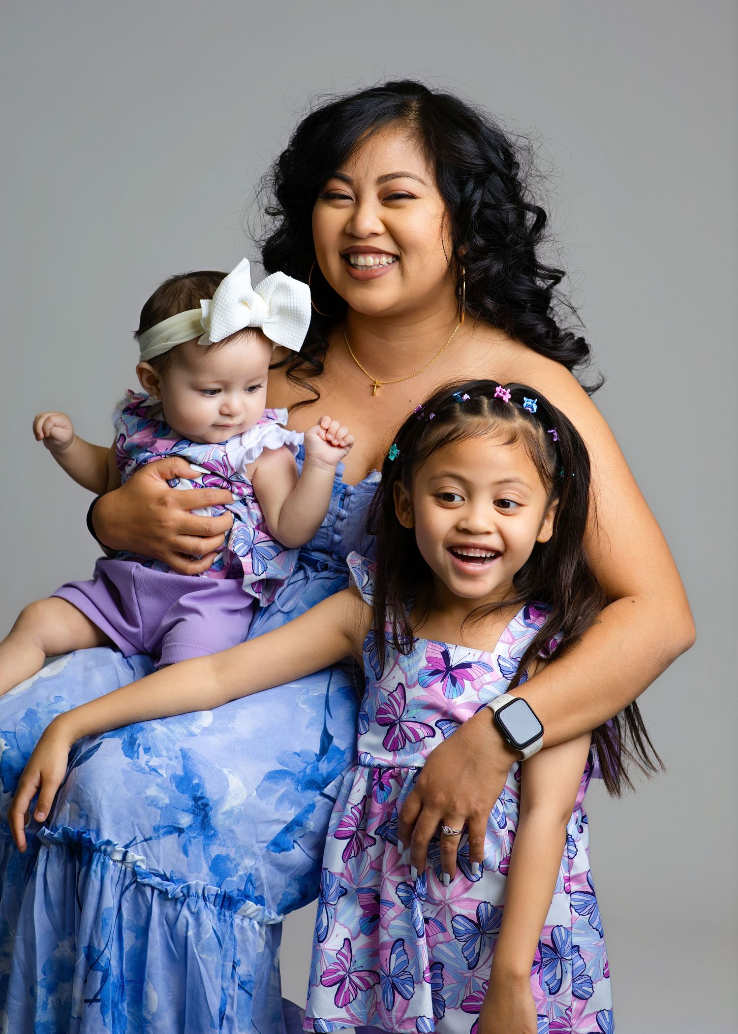 Smiling mother in blue floral dress holding baby and young daughter in butterfly dress.