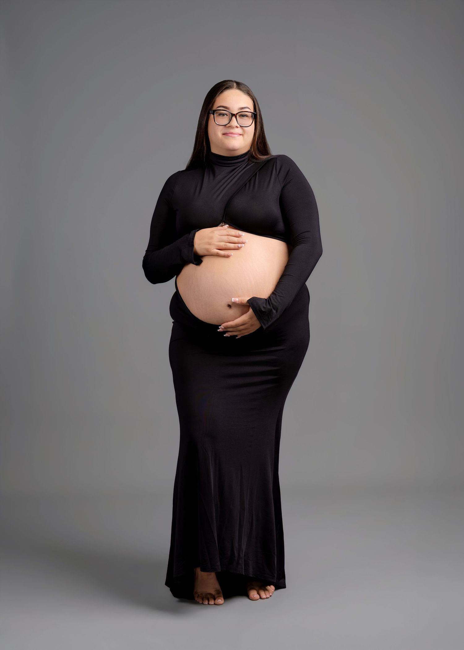 Pregnant woman in glasses and a fitted black long-sleeve maternity outfit stands barefoot, cradling her bare belly in a studio portrait.