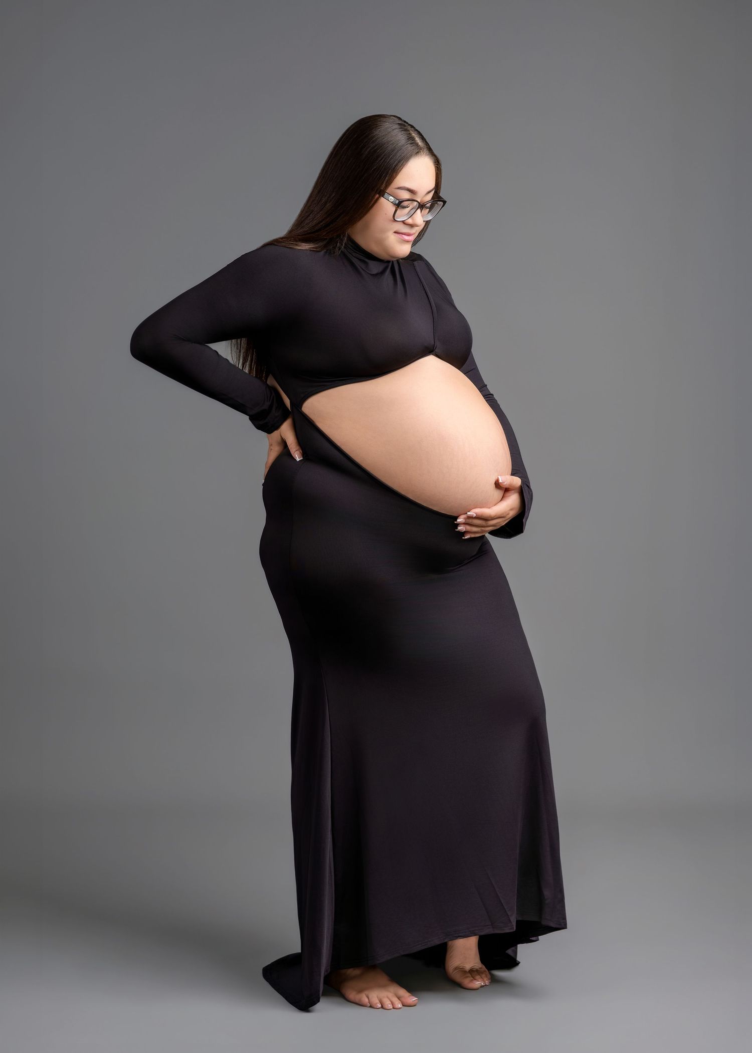 Pregnant woman in black cutout maternity gown cradles baby bump during professional studio maternity photoshoot.