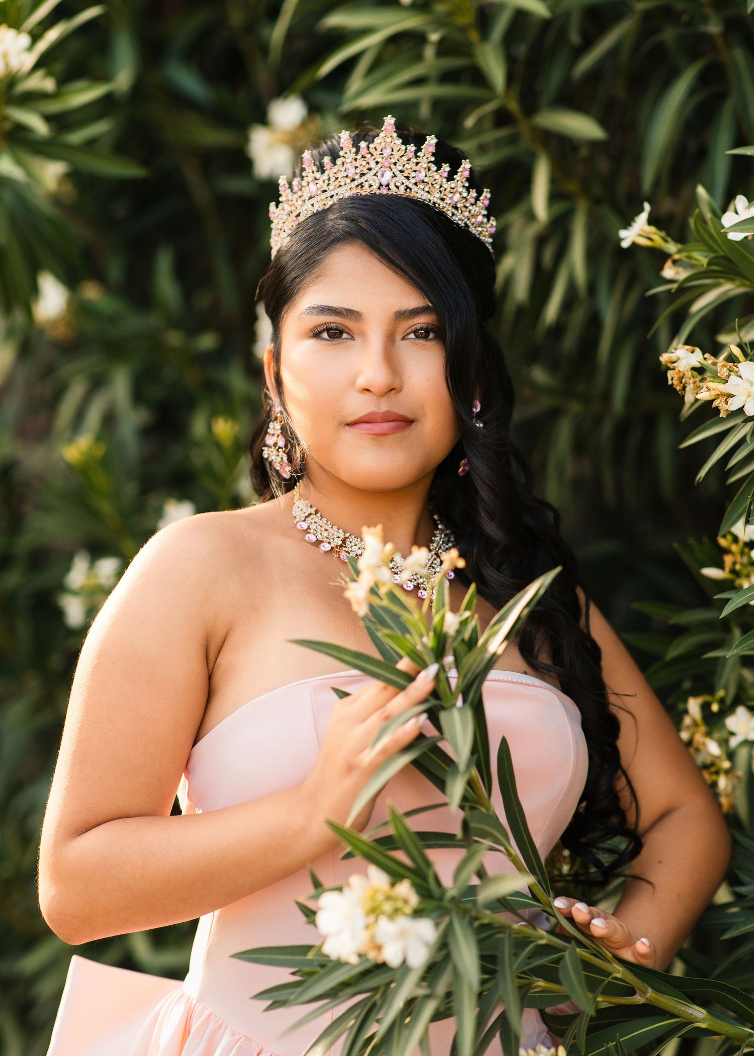 Young Latina woman in pink strapless gown wearing a gold tiara poses among white flowering bushes.