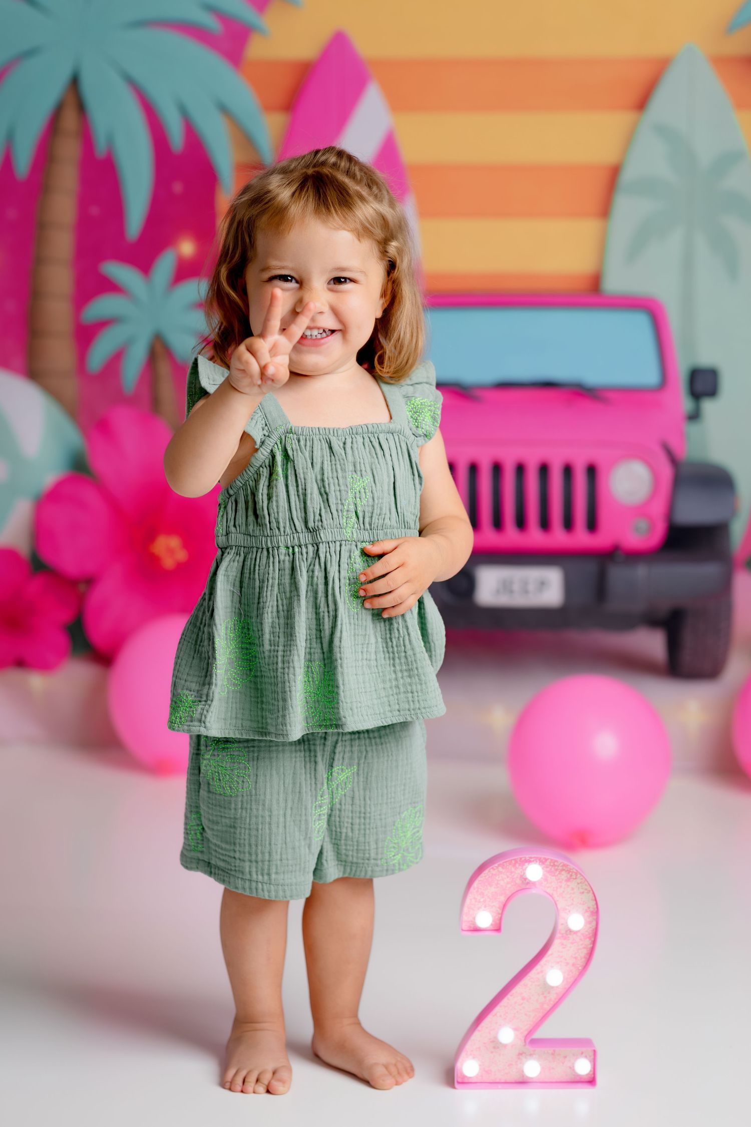 Smiling toddler girl in green outfit holding up two fingers at a tropical birthday party.