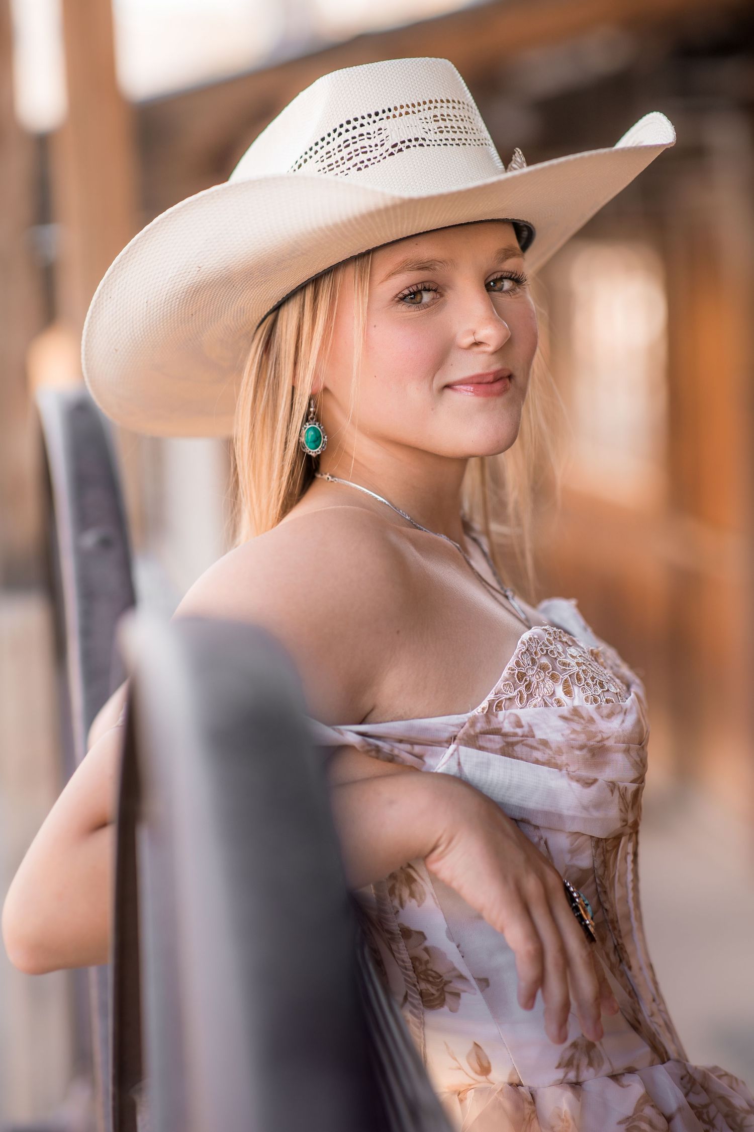 Blonde woman in floral dress and white cowboy hat poses for senior portrait at rustic outdoor venue.