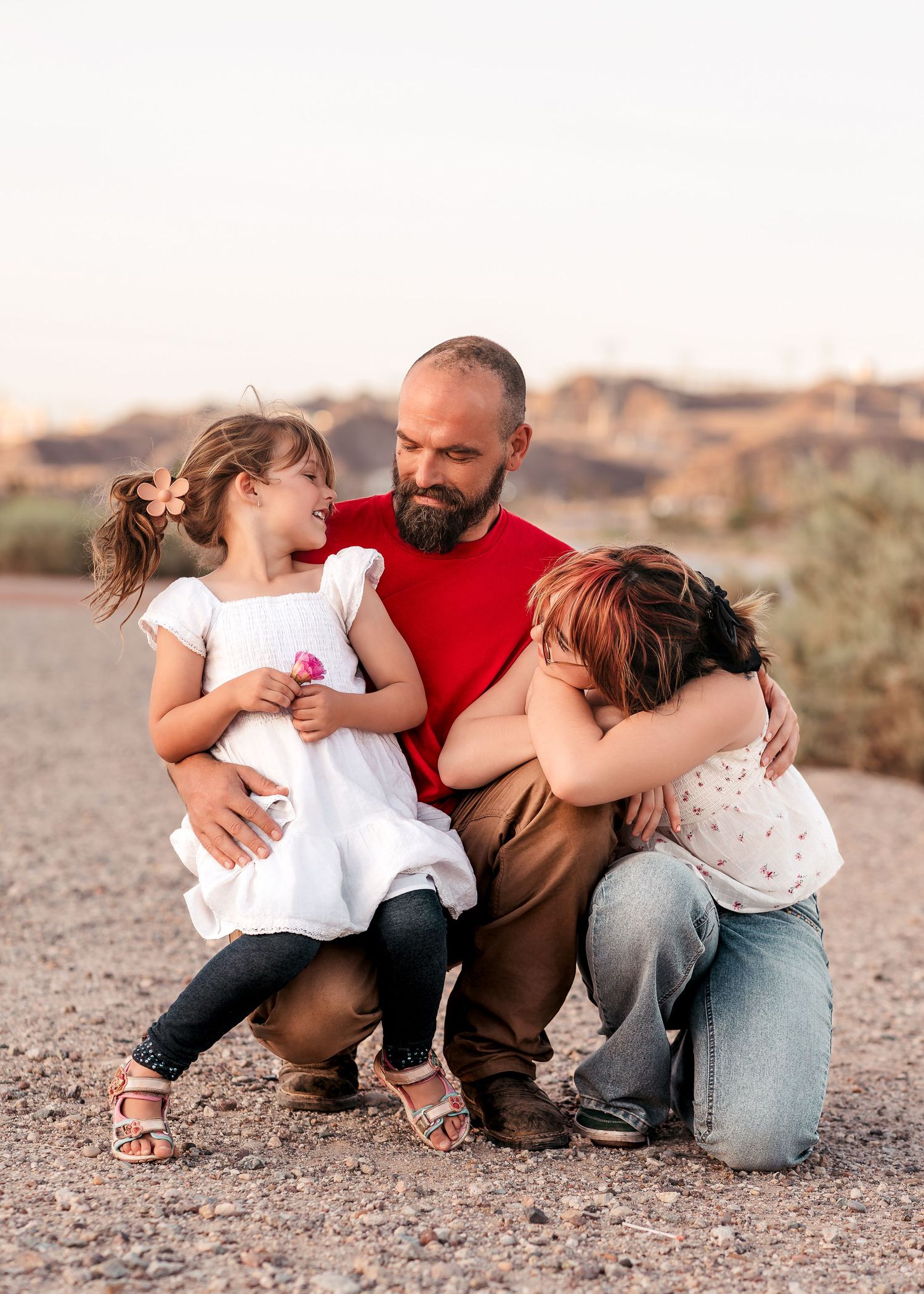 A bearded father crouches outdoors hugging two young daughters in a desert landscape at golden hour.