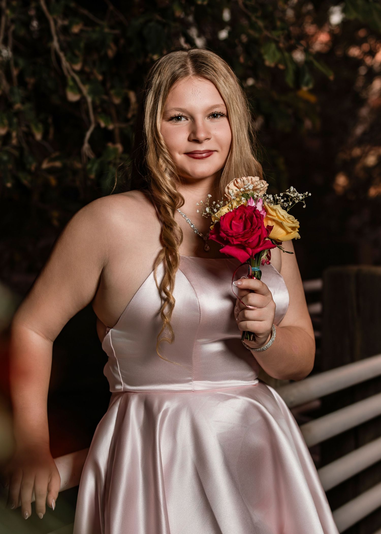 Young woman in pink satin dress holds red and yellow roses at outdoor evening event.