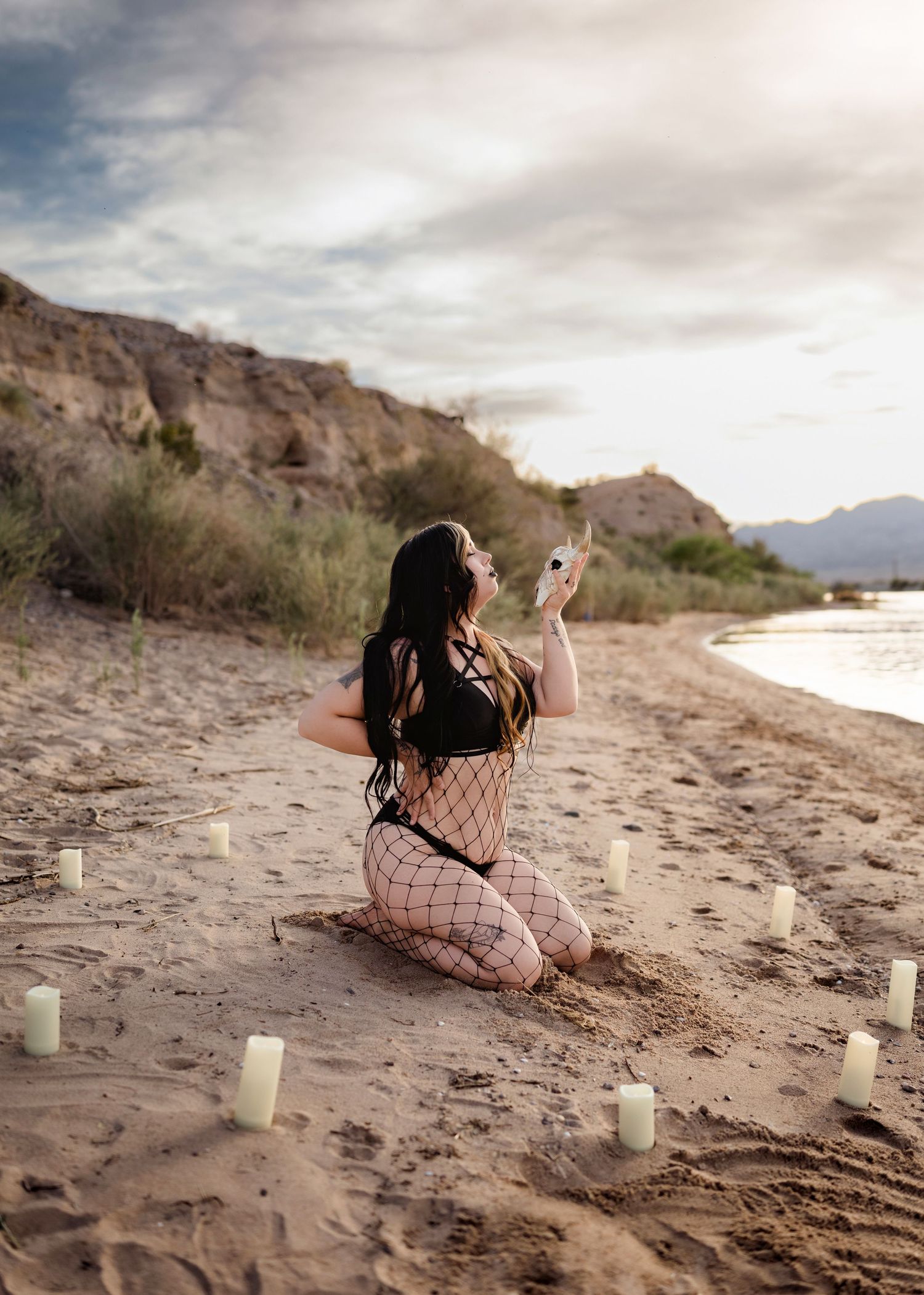 Woman in fishnet and black outfit kneeling on sandy beach with white candles at sunset.
