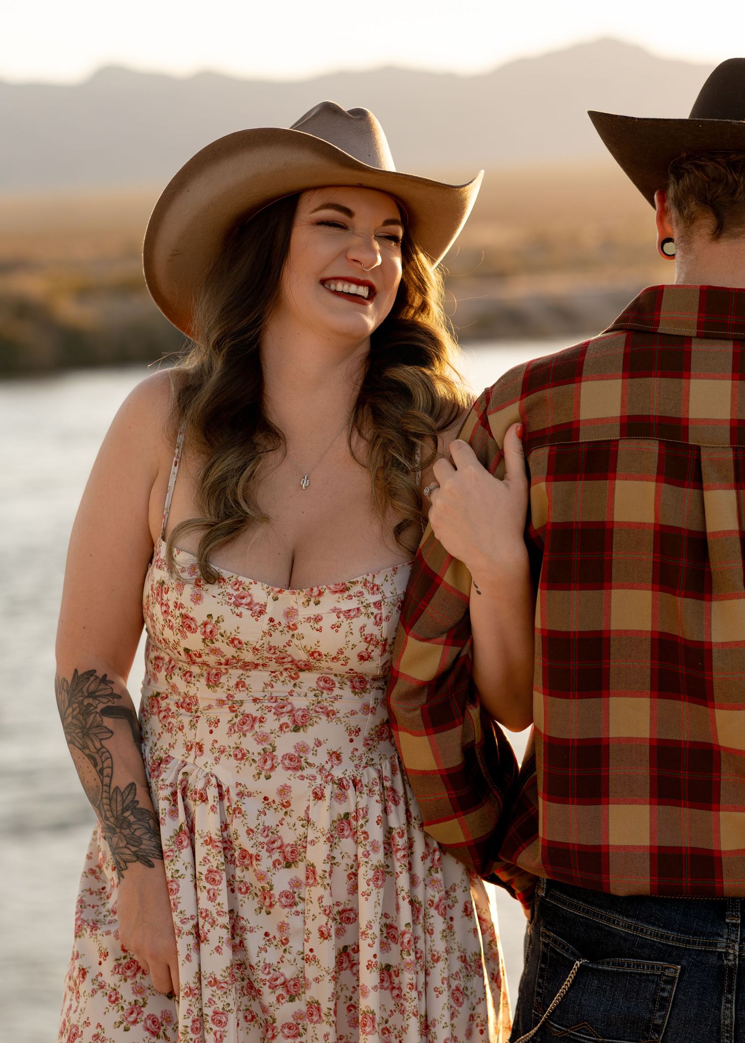 A laughing woman in a floral sundress and cowboy hat enjoys an outdoor moment near water.