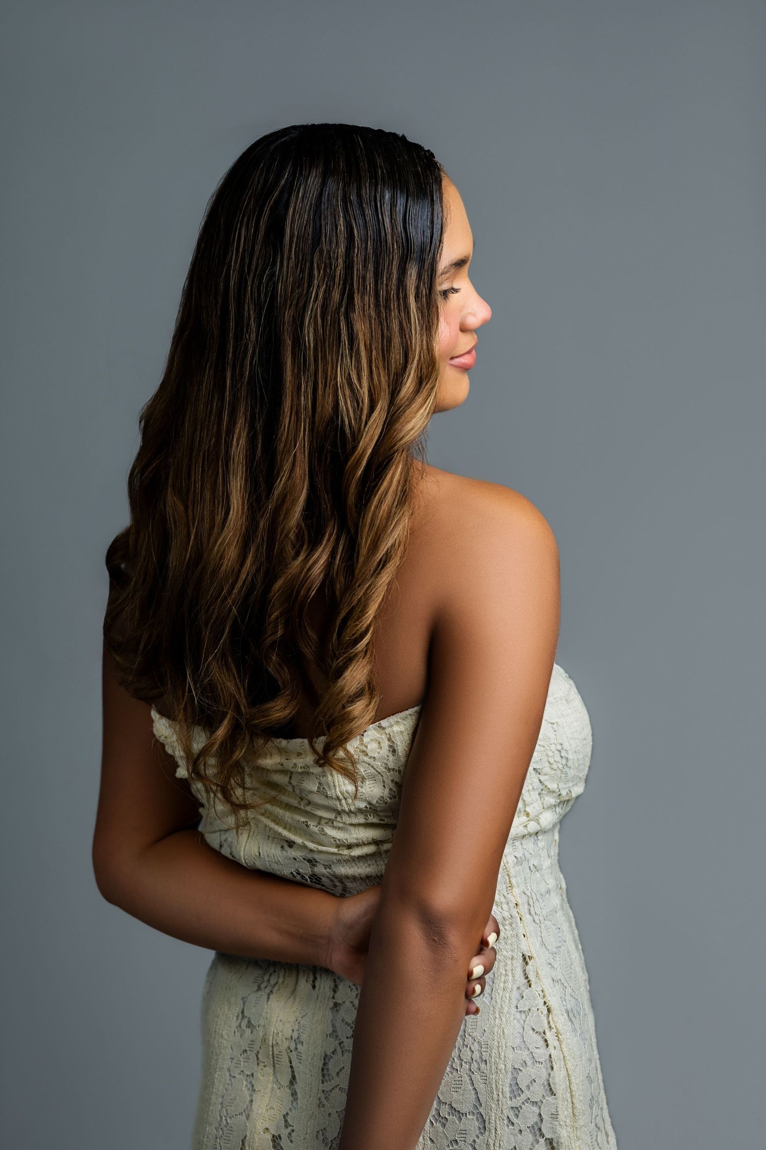 A young woman with long wavy highlighted hair poses in a white strapless dress, side profile view.