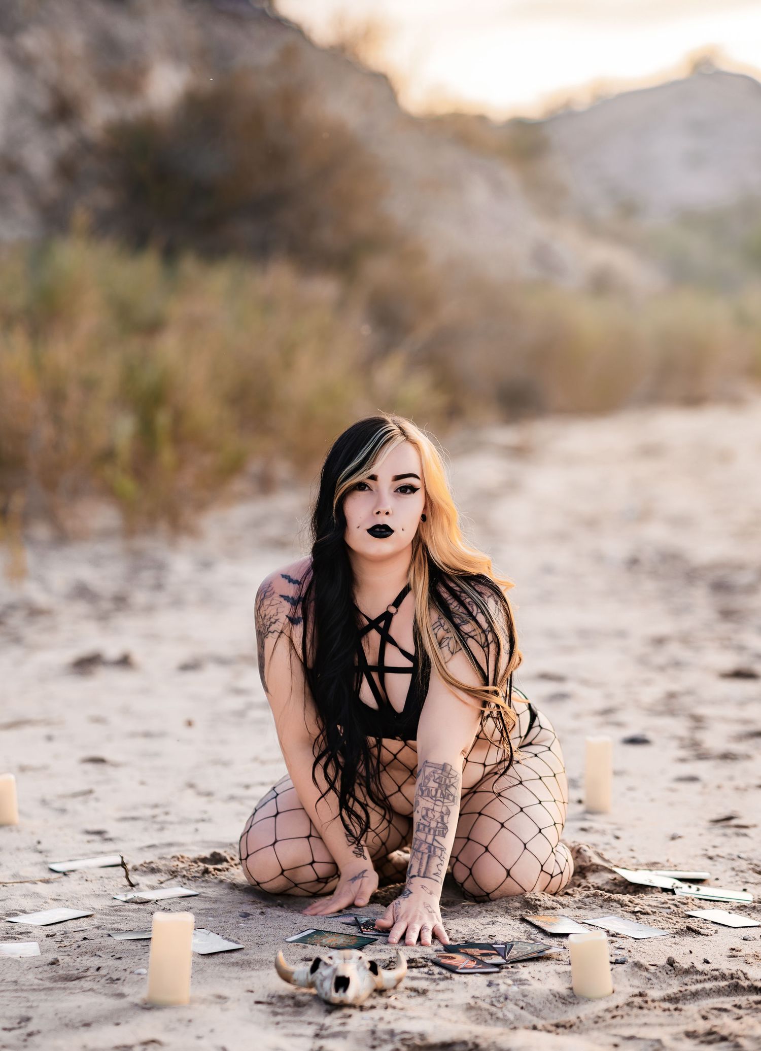 Tattooed woman in gothic black outfit with tarot cards and candles posed on sandy desert ground.