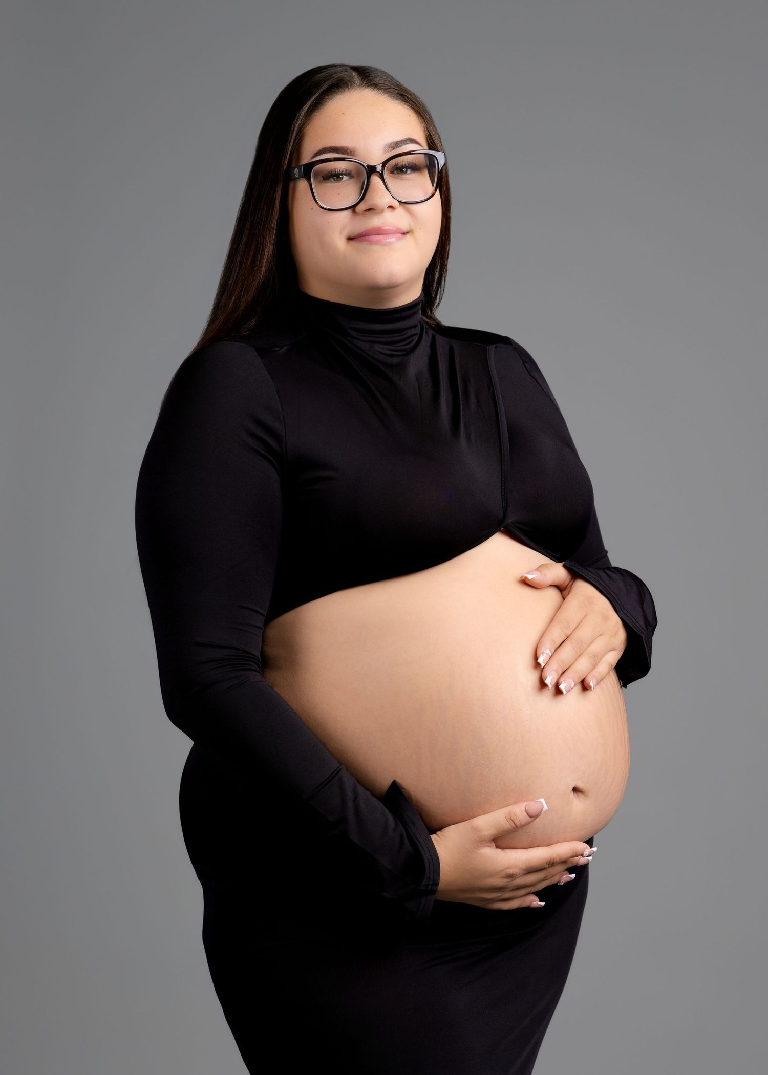 A pregnant woman in a black turtleneck dress holds her bare baby bump in a studio portrait.