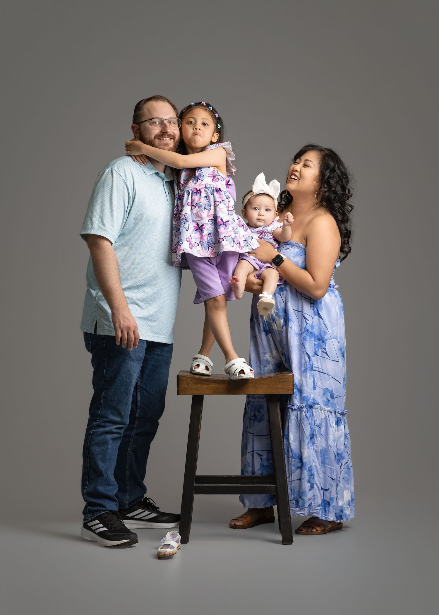 Smiling family portrait of a father, mother, young girl standing on a stool and baby in a bow headband against a gray studio backdrop.
